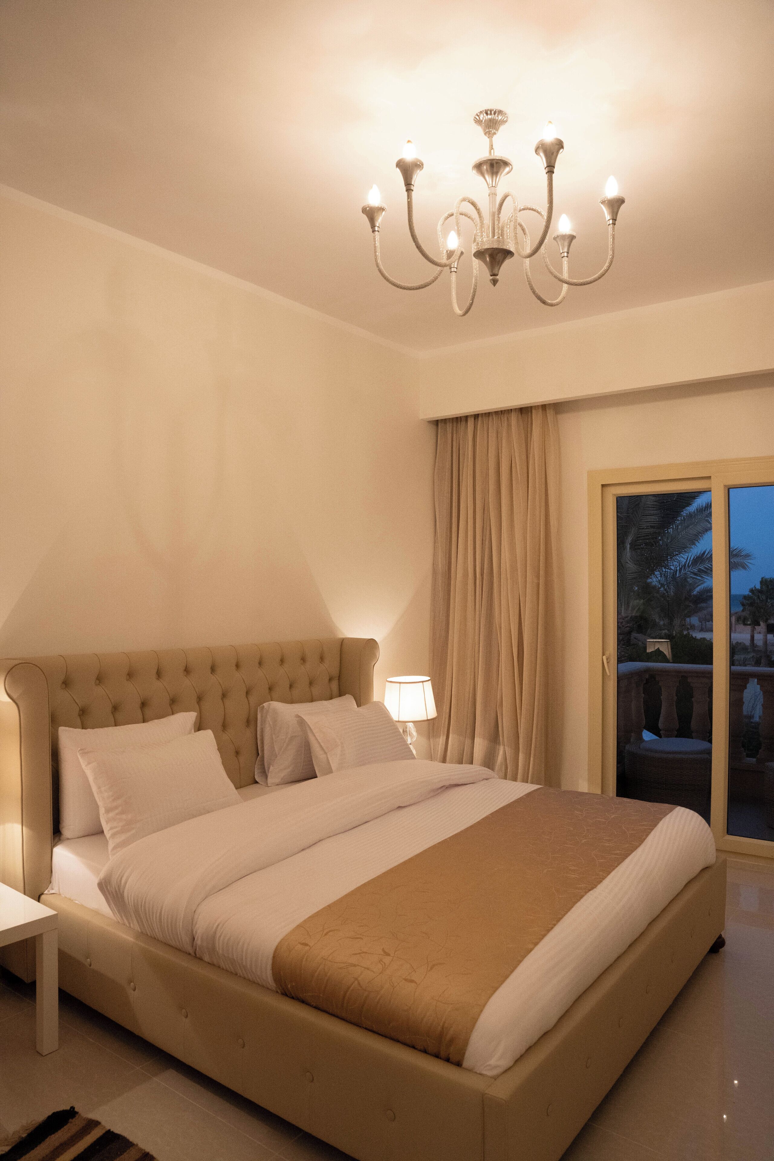 Ein Schlafzimmer mit einem großen Bett, einer Kandelaber, einem Nachttisch und Blick auf Palmen durch eine Schiebetür.