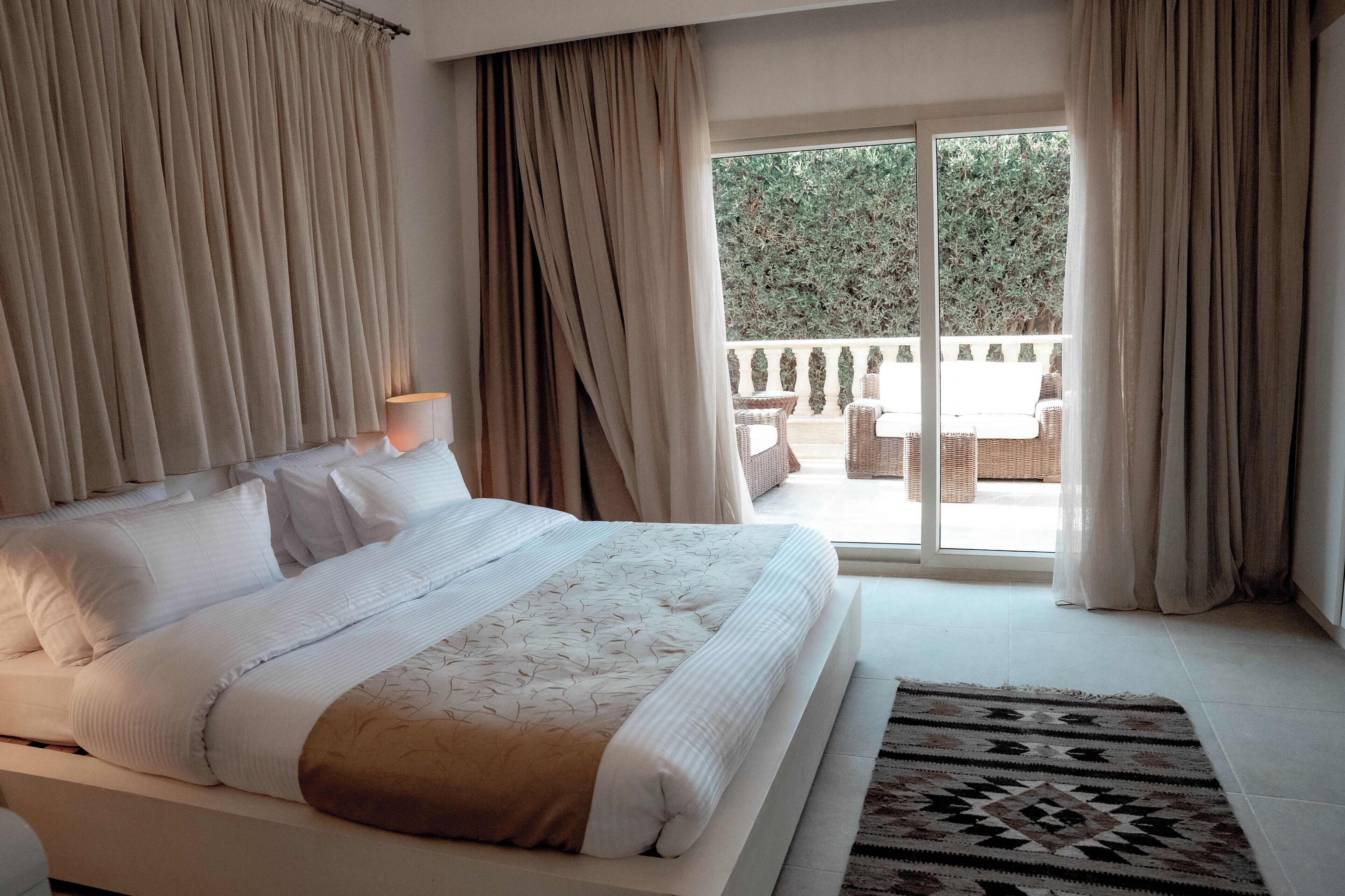 A bedroom with a large bed, a view of the outdoors, and a balcony with furniture.