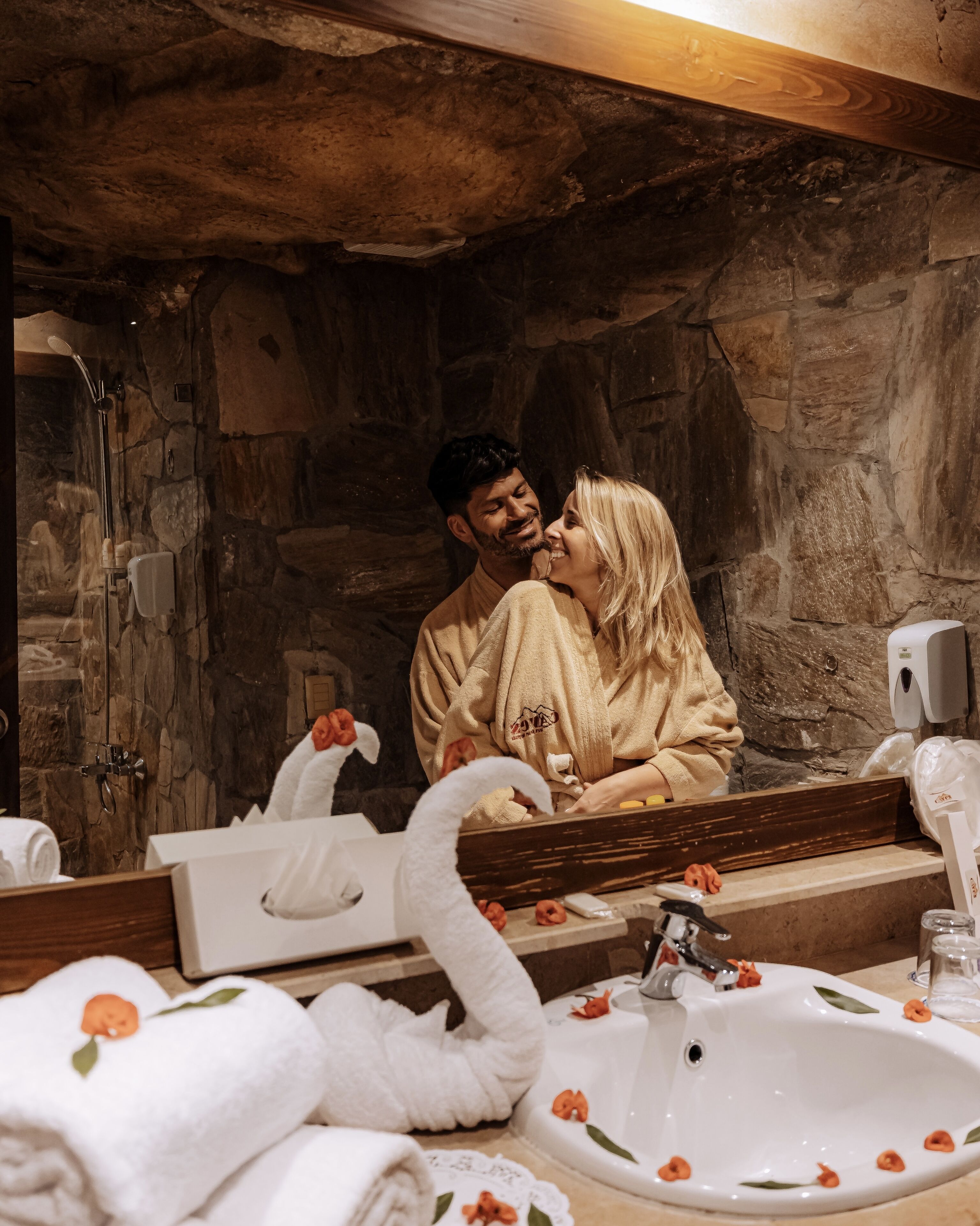 A stone-walled bathroom with a wooden vanity, a large mirror, and a white sink with decorative flowers.