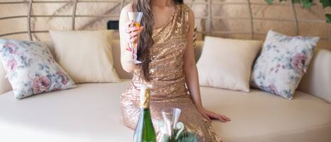 Una mujer con un vestido dorado sentada en un sofá blanco con cojines estampados, sosteniendo una copa y una botella.