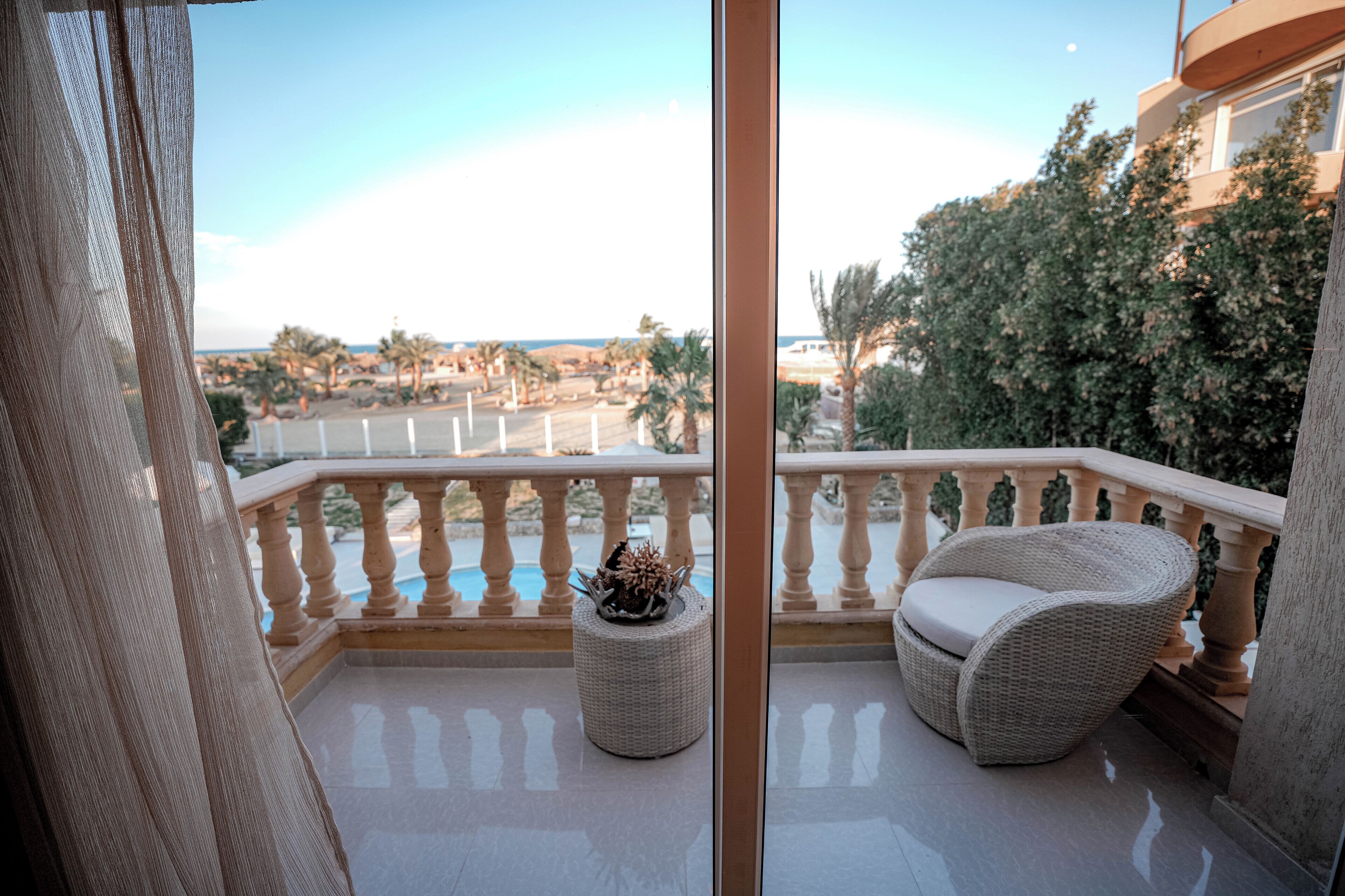 A balcony with a wicker chair and ottoman, a pool, and a view of a residential area with trees and buildings.