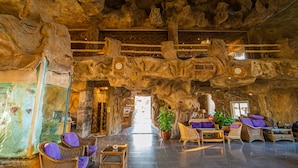 Een ruime lobby met een hoge plafond en natuurlijke rotsformaties. Er staan rieten stoelen en een grote potplant.