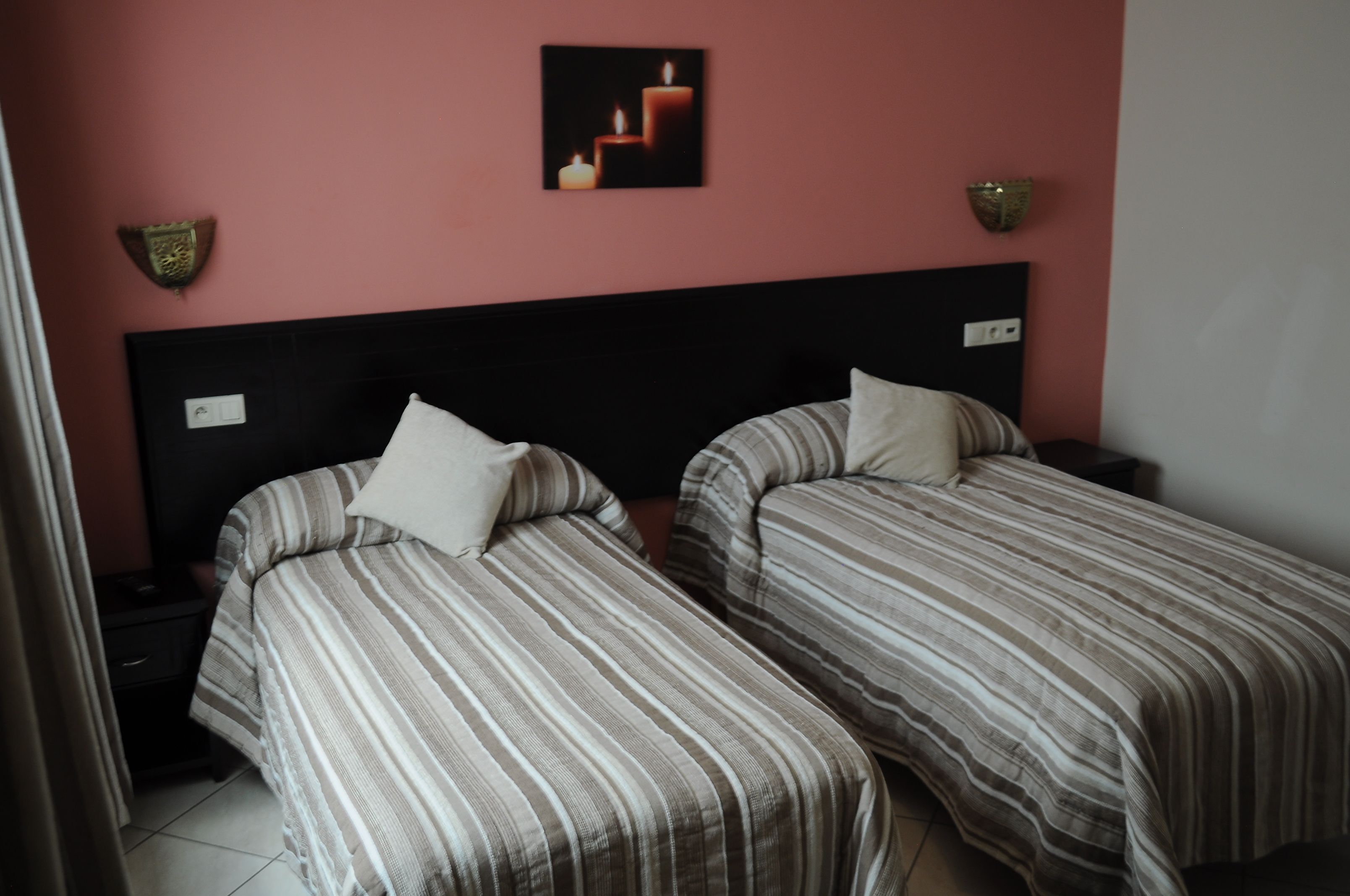 A room with two single beds, a black headboard, a nightstand with a lamp, and a wall-mounted picture of candles.