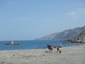 Een rotsachtig strand met mensen en een vuurtoren in de verte.