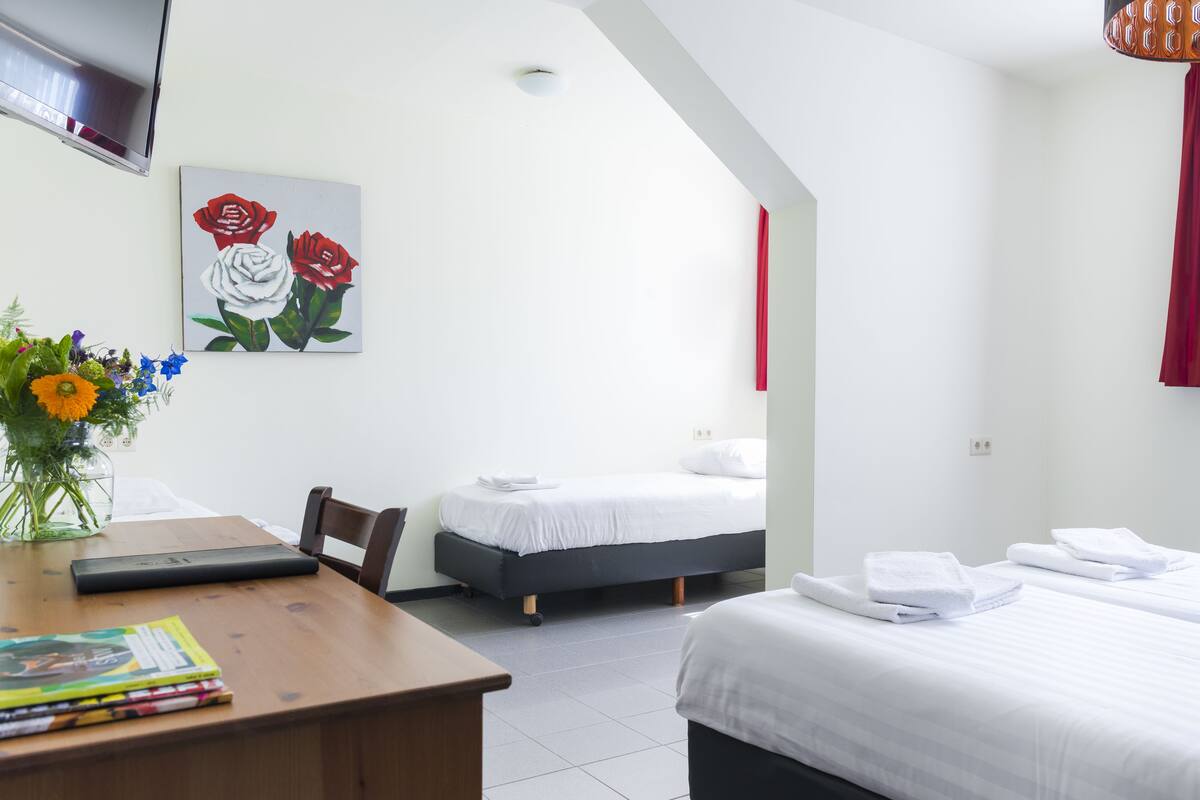 A hotel room with a bed, a desk with a laptop, a vase with flowers, and a painting of roses on the wall.