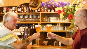 Two people toasting with drinks at a bar with shelves of bottles and glasses behind them. - Hotel De Kruishoeve (Vught)
