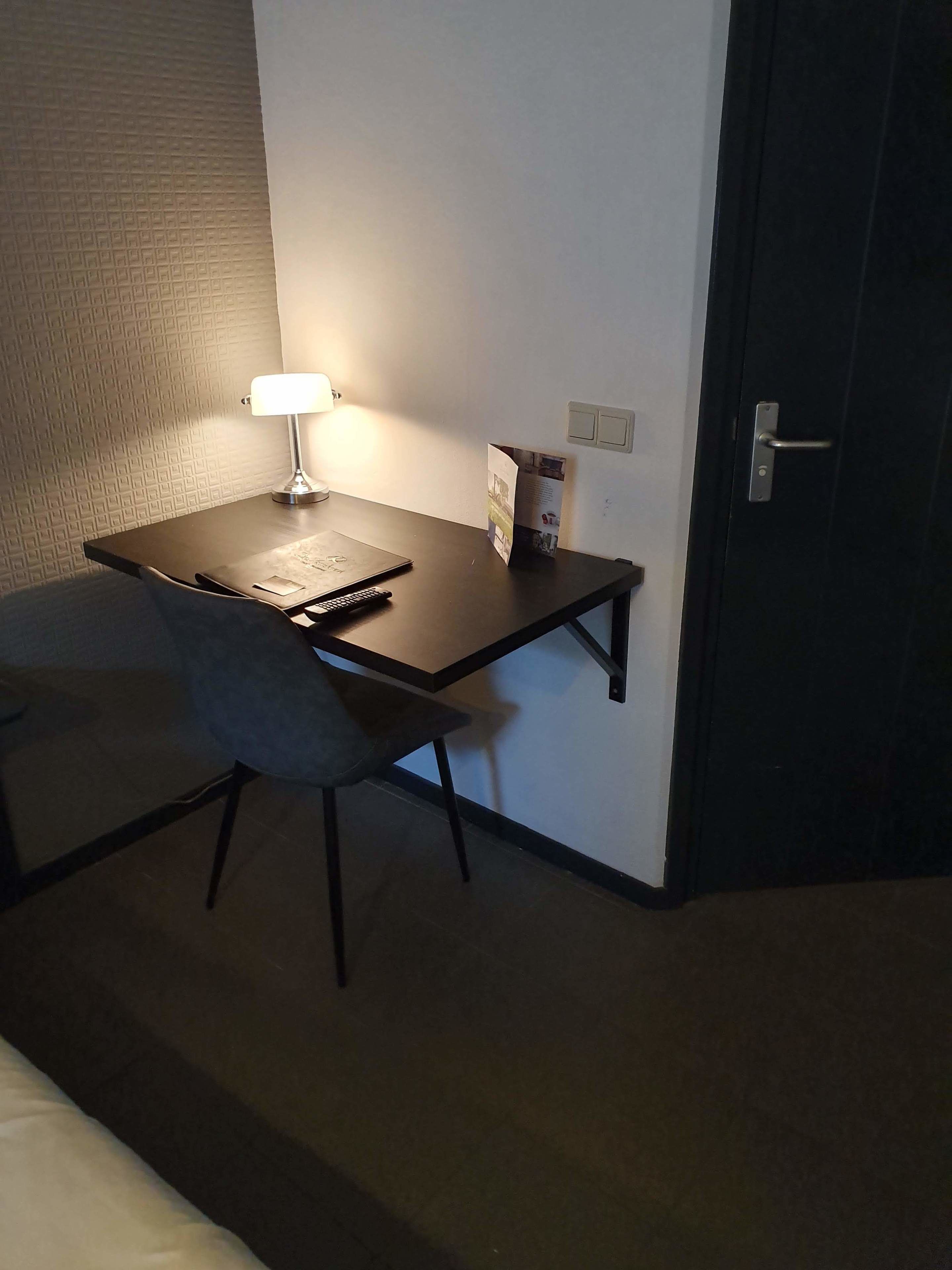 Une petite pièce sombre, avec un bureau, une chaise, une lampe et une porte.