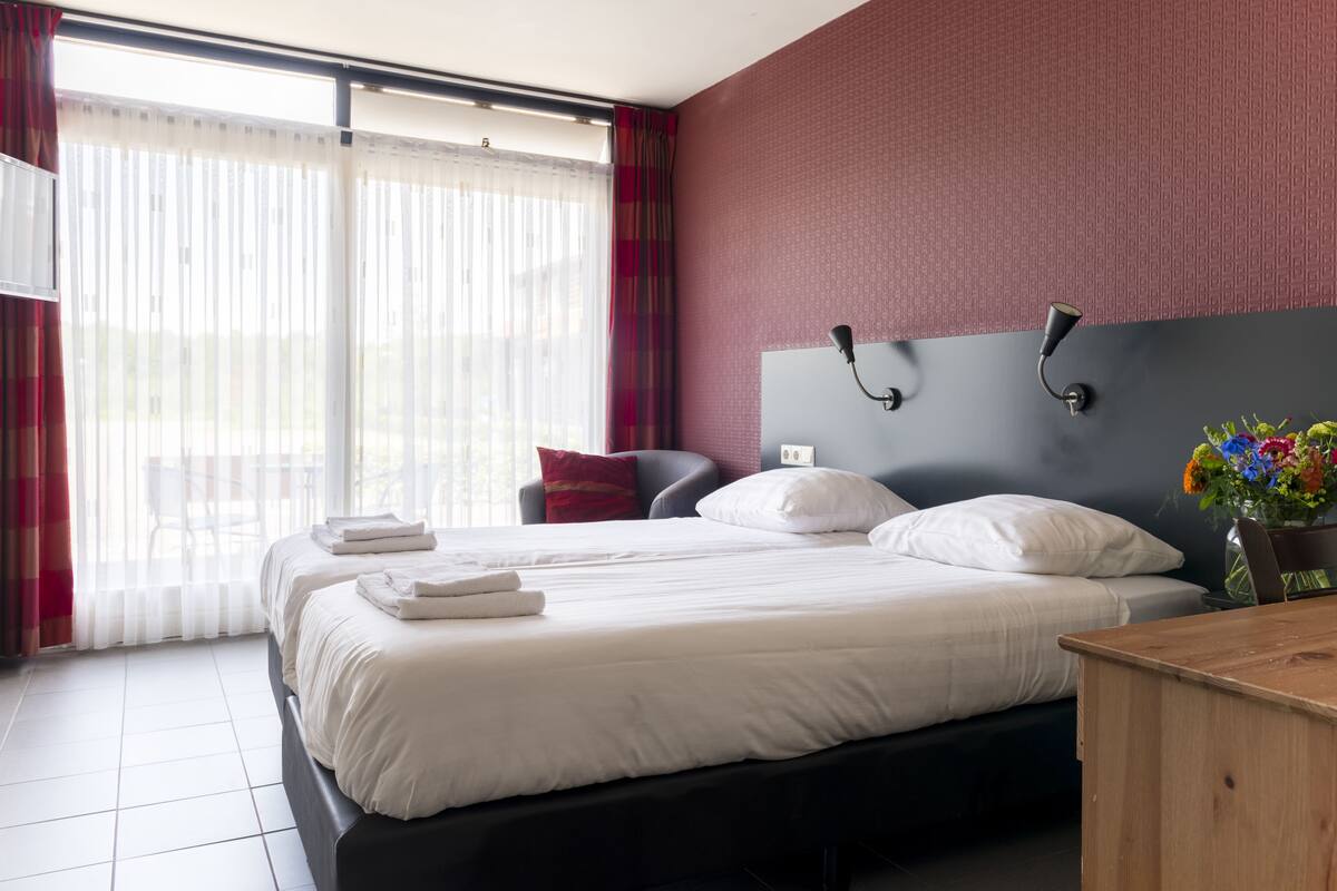 A hotel room with two beds, a large window with sheer curtains, a red textured wall, a black headboard, and a wooden nightstand with a vase of flowers.