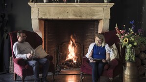 Two people seated on red sofas in a room with a fireplace, a vase of flowers, and a metal container on the mantelpiece. - Hotel De Kruishoeve (Vught)