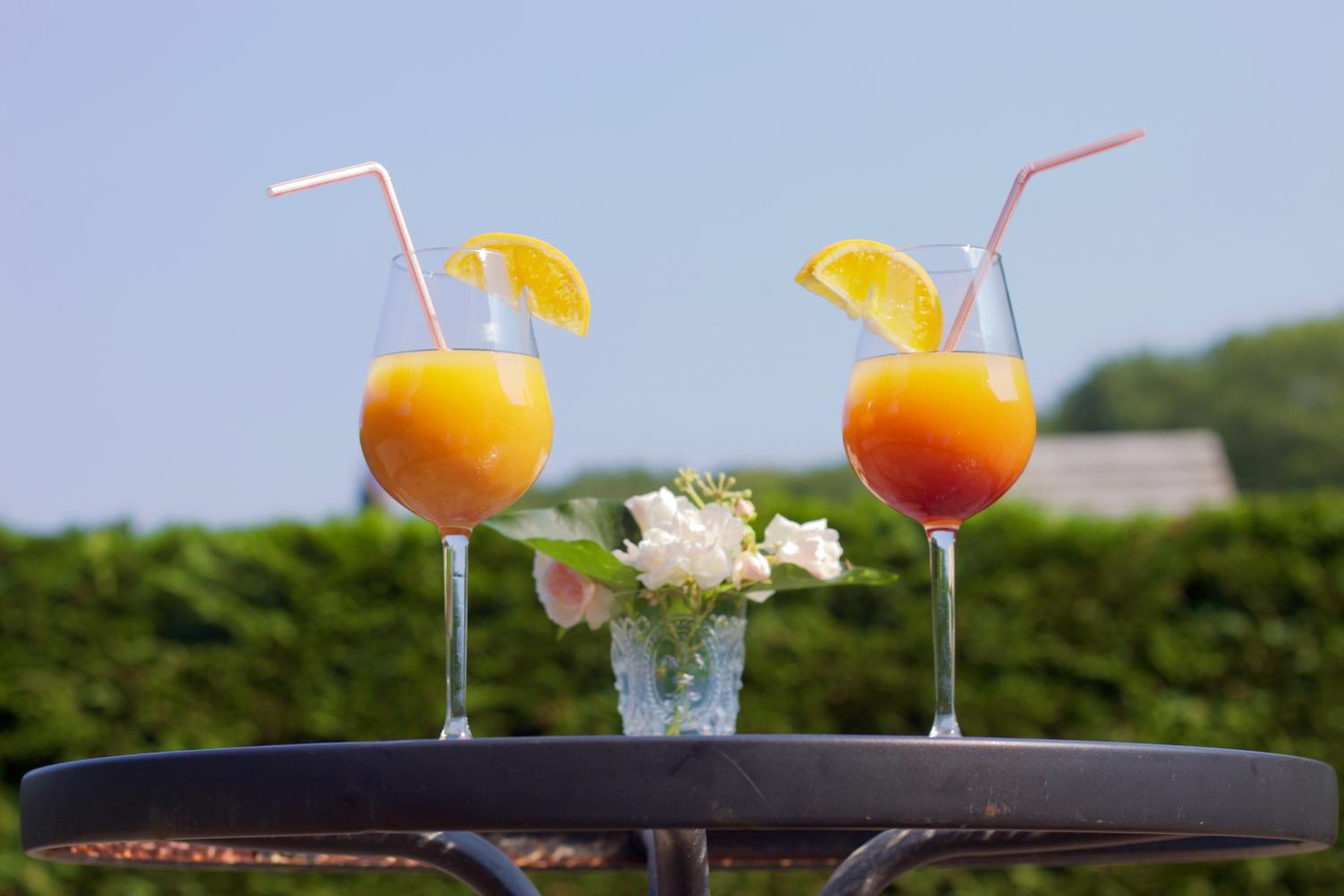Two glasses of orange juice on a table with a clear sky and greenery in the background.