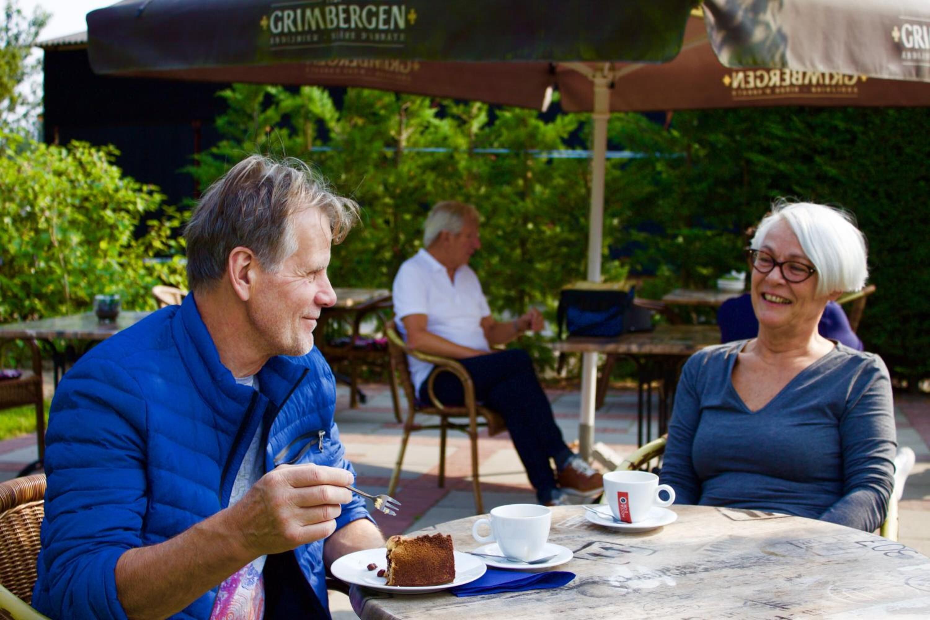 Two people sitting at an outdoor table with a cake and a cup.