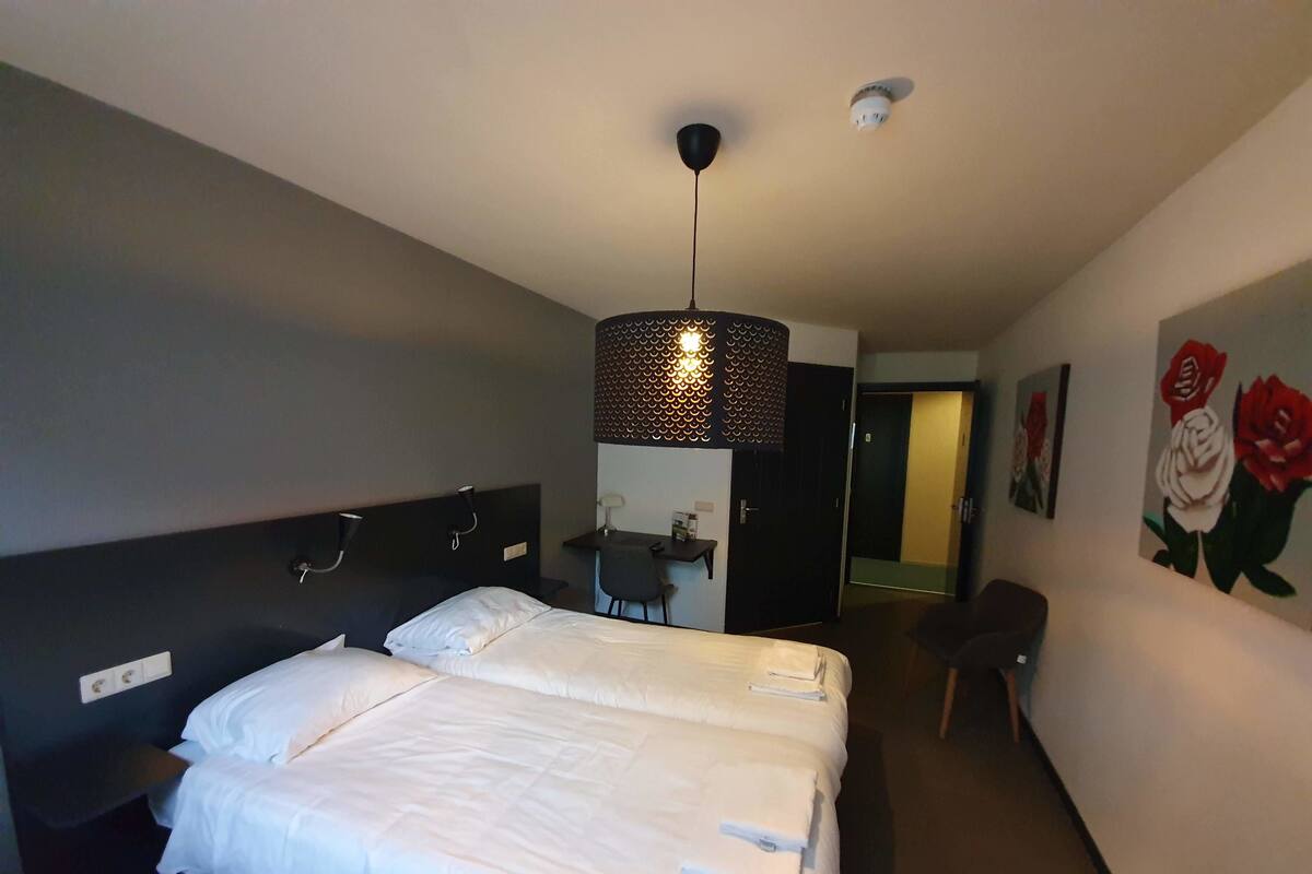 A hotel room with a large bed, a desk, and a chair. There is a painting of roses on the wall.