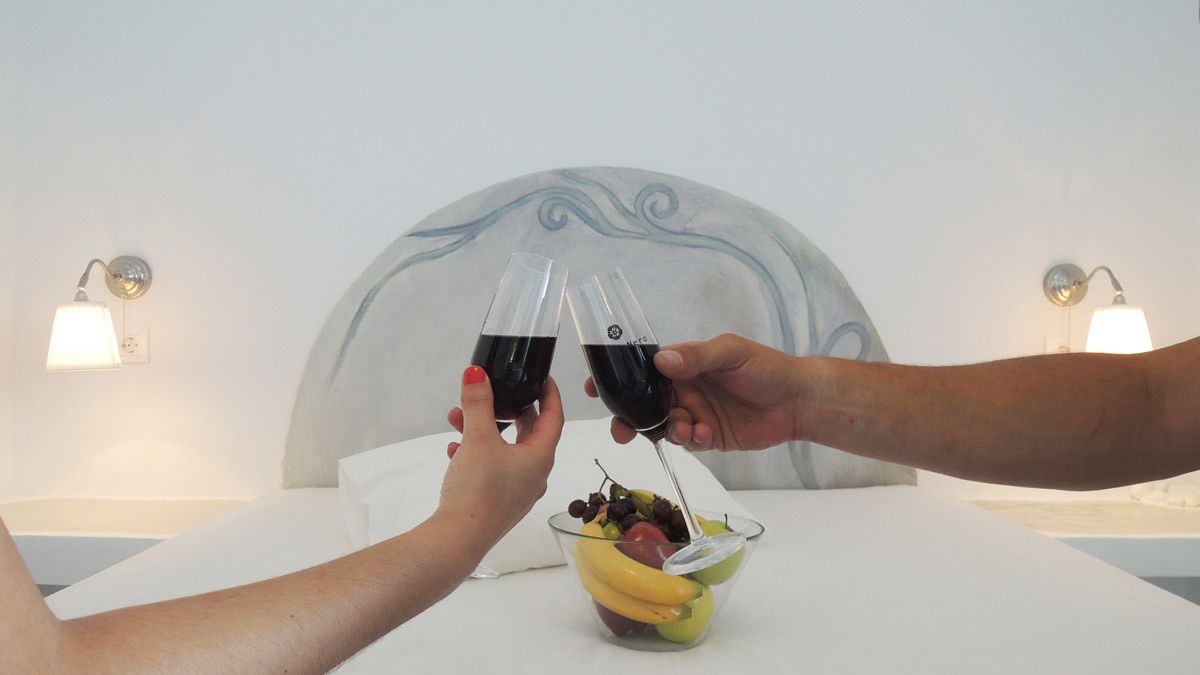 Una persona brindando a otra con una copa de vino tinto, con un bol de fruta y una mesa blanca al frente.