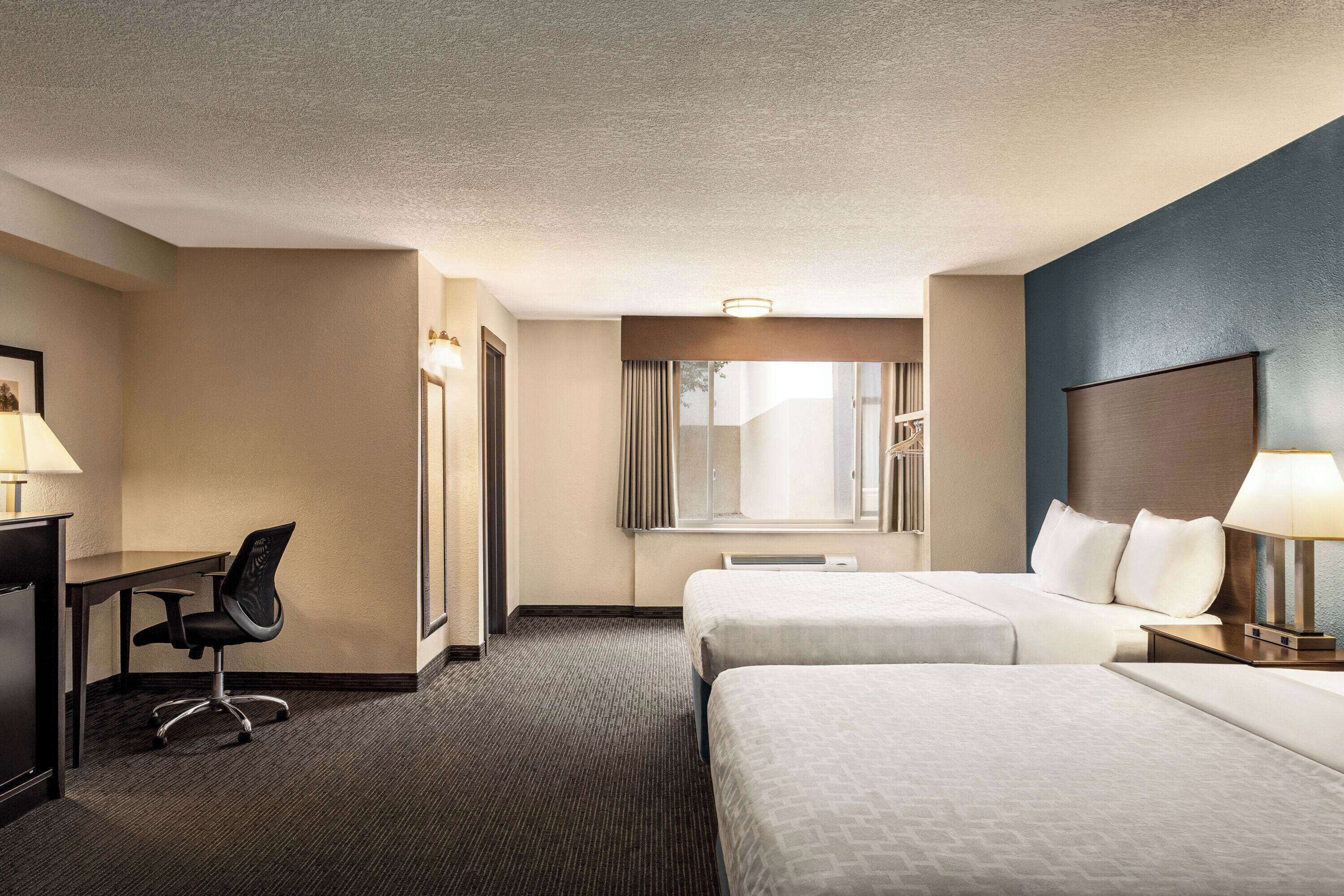 Une chambre d’hôtel avec deux lits, un bureau, une chaise, une fenêtre et une lampe.