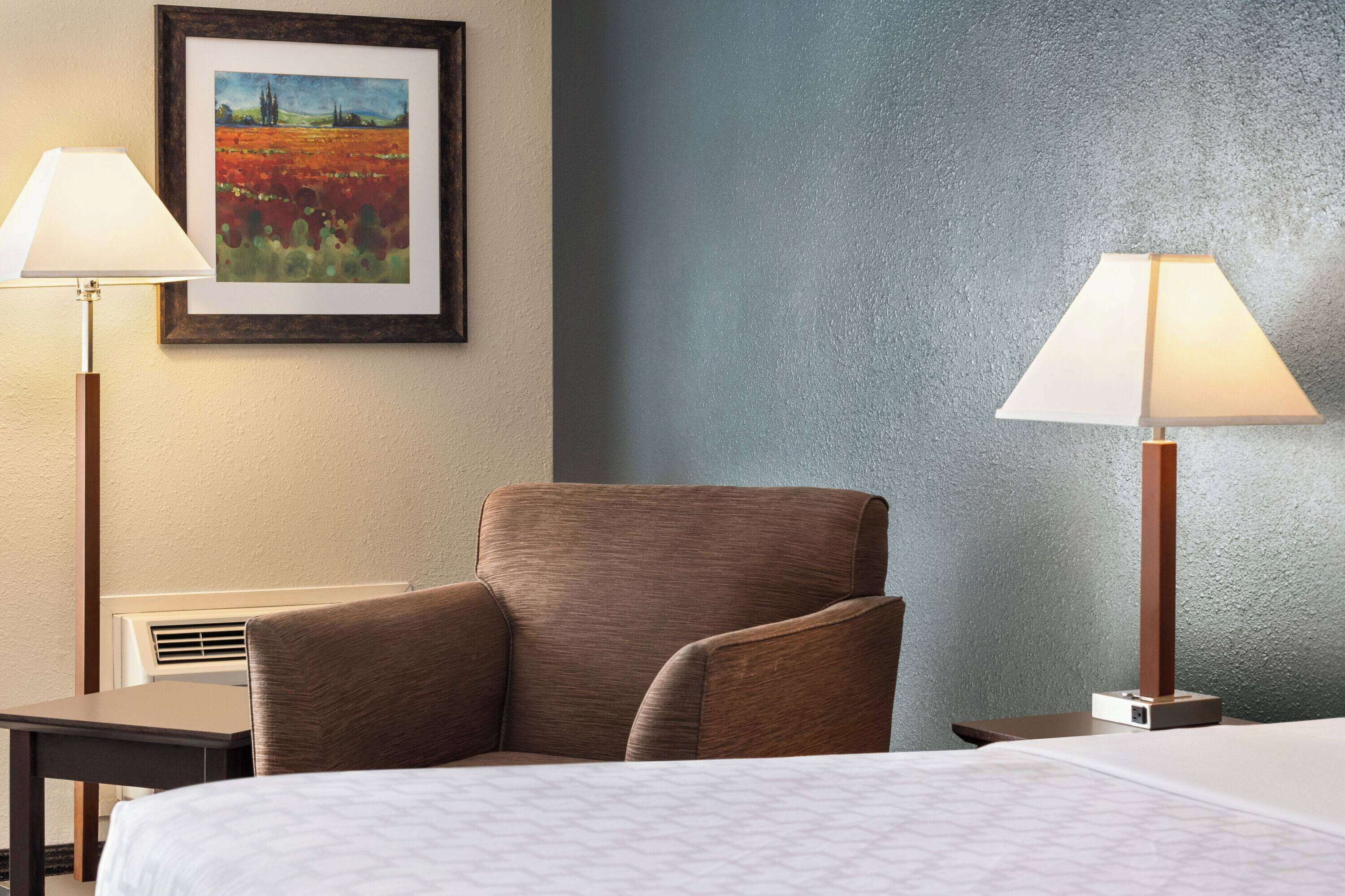 A hotel room with a bed, two lamps, a chair, a desk, and a framed painting on the wall.