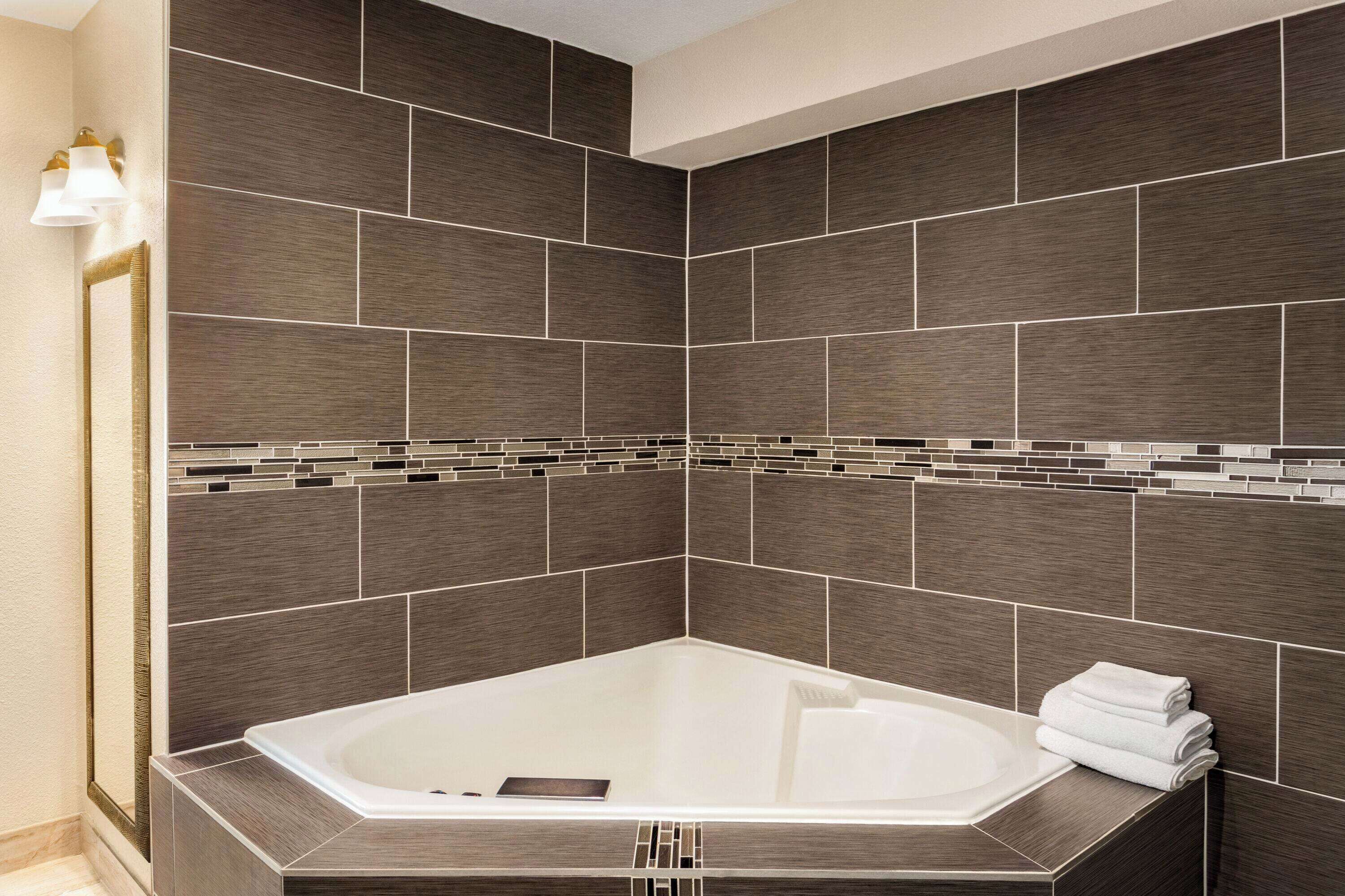 A modern bathroom with a large bathtub, dark tiled walls, and a small mirror with a light fixture.