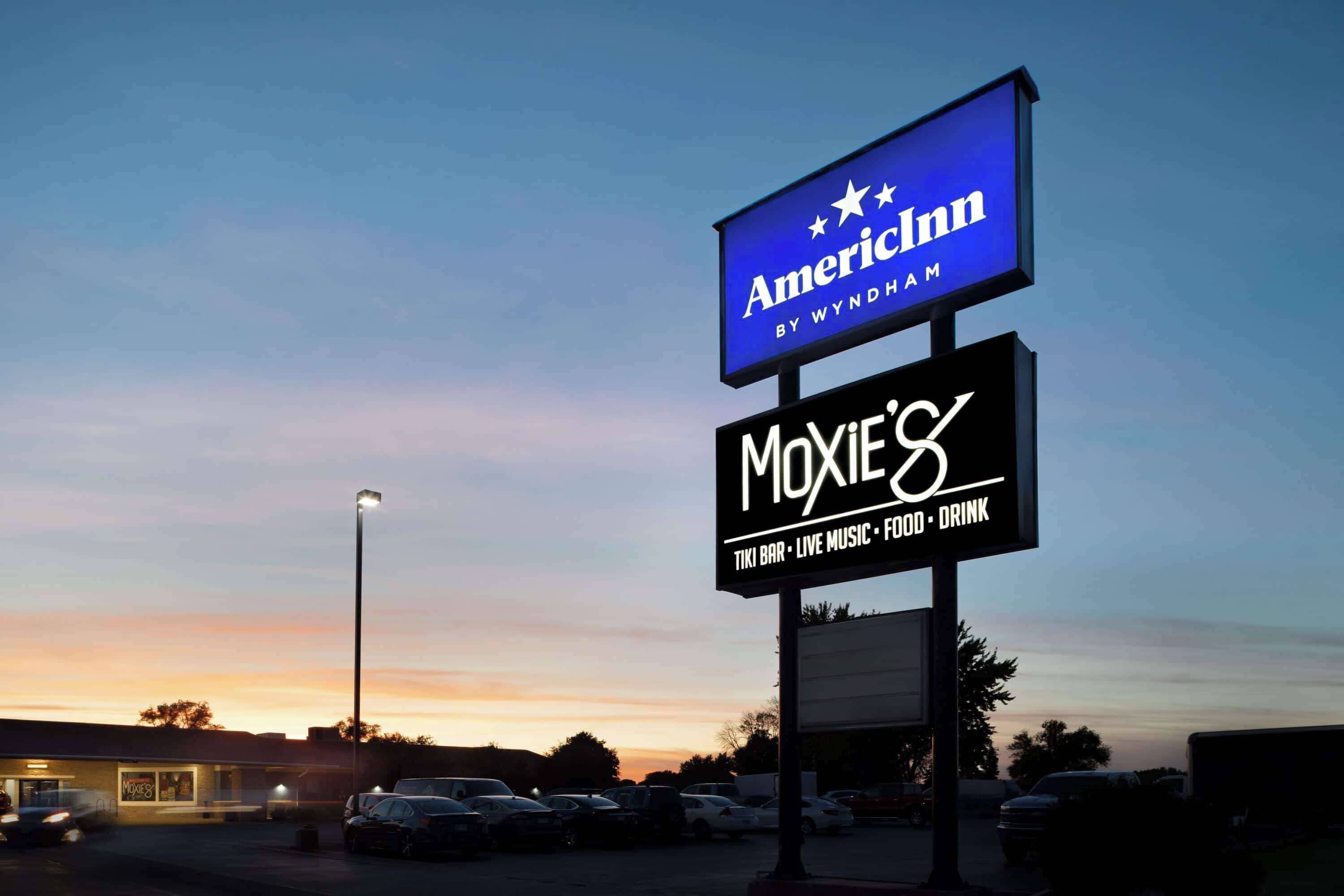 Un estacionamiento con un cartel que indica 'American Inn by Wyndham' y 'Moxie's', que ofrece un bar tiki, música en vivo, comida y bebidas.