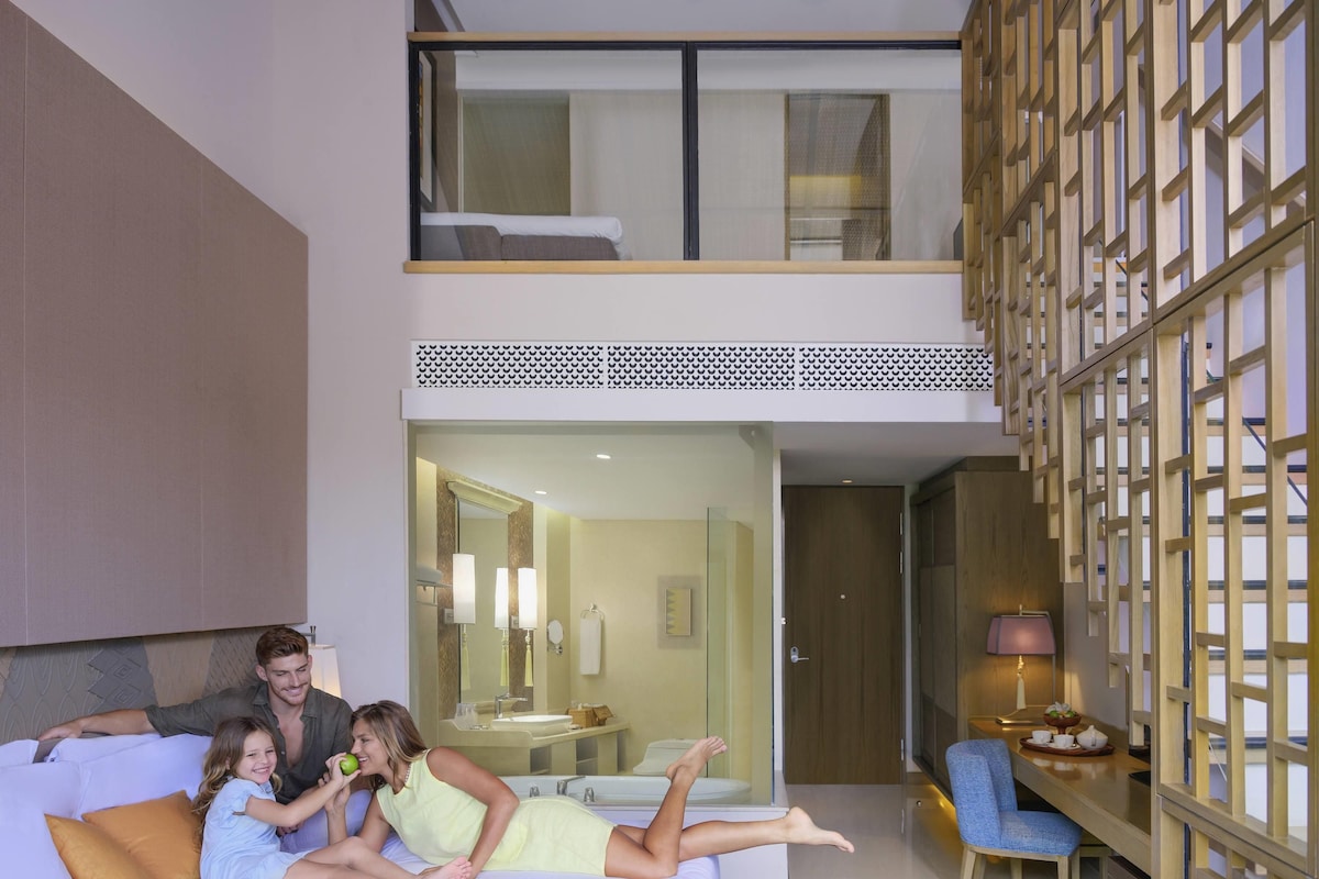 A modern hotel room with a bed, a seating area with a chair, a small table, and a view of the bathroom.