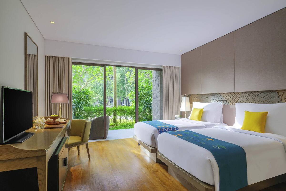 A hotel room with a large bed, a desk with a TV, and a view of greenery through the window.