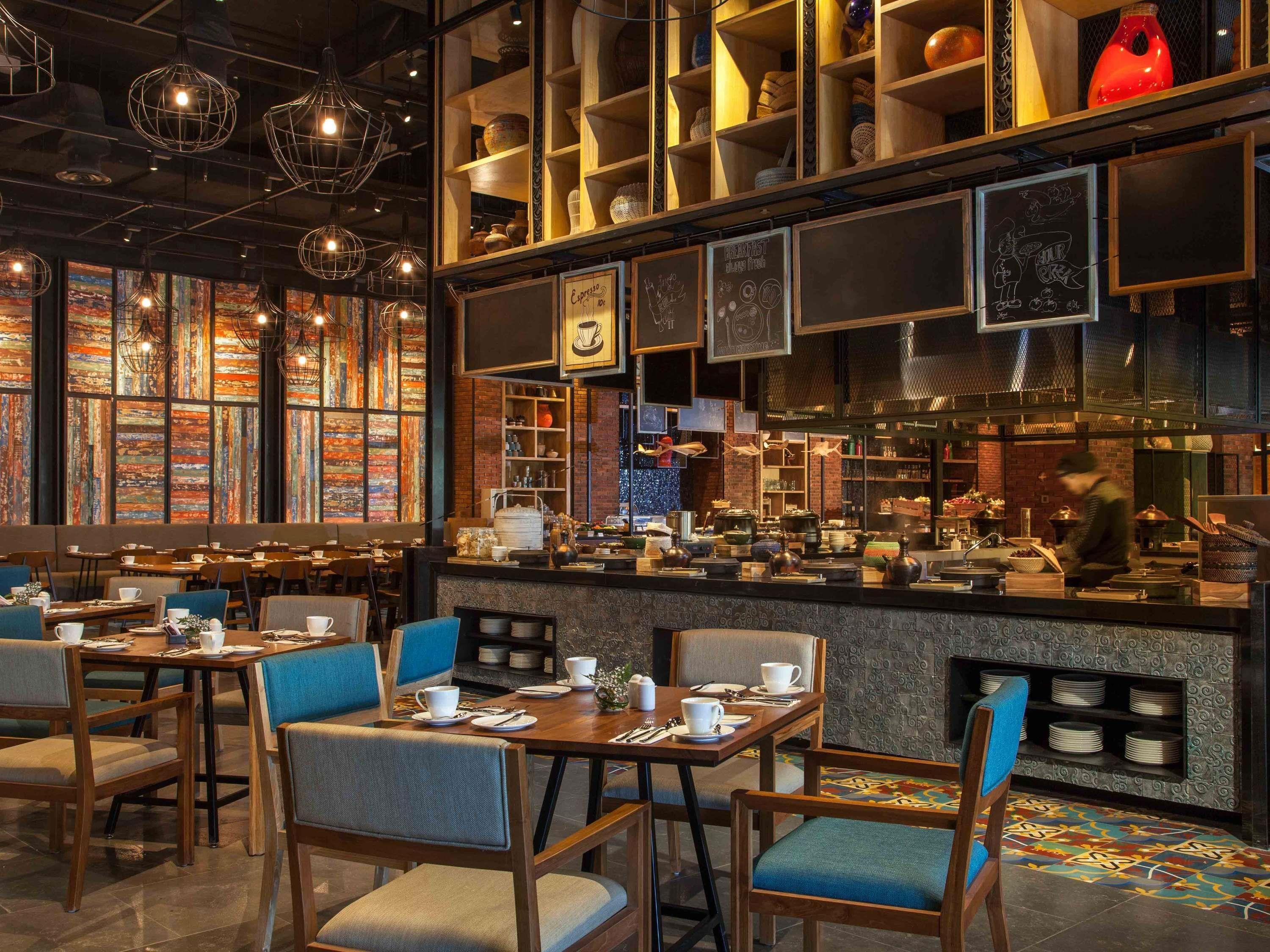 A restaurant with wooden tables and chairs, a bar area with a chef, and shelves with various kitchen items.