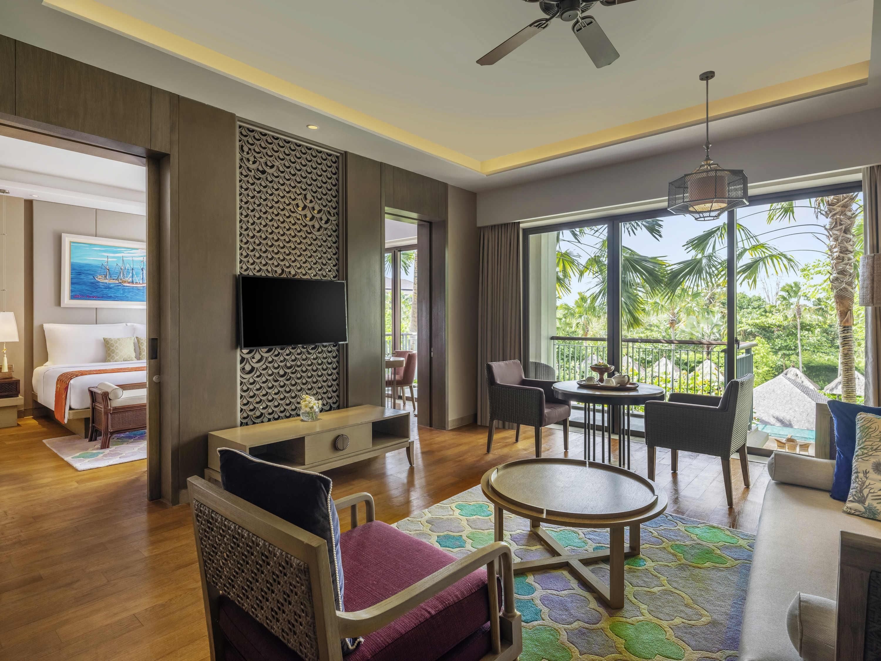 A modern hotel room with a bed, a TV, a seating area with a table, and large windows offering a view of palm trees.