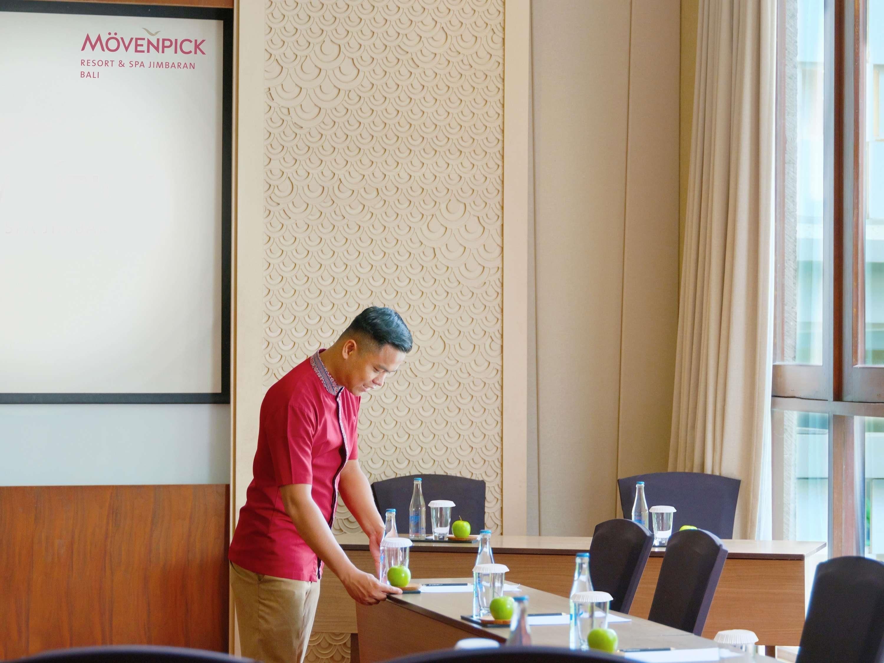 A man in a red shirt arranging items on a table in a room with a patterned wall and a large window.