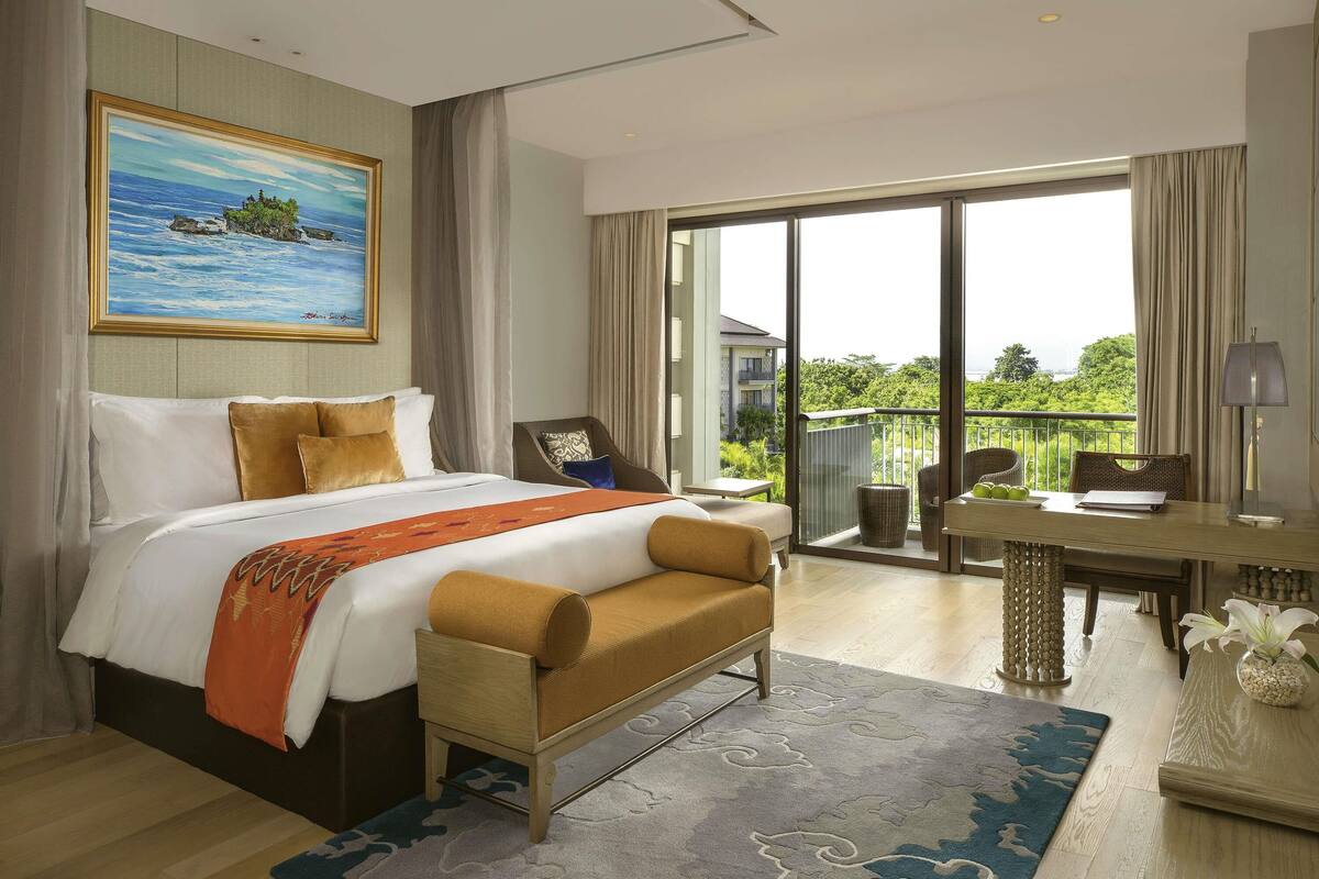 A hotel room with a bed, a desk, a chair, a sofa, and a balcony with a view of greenery.