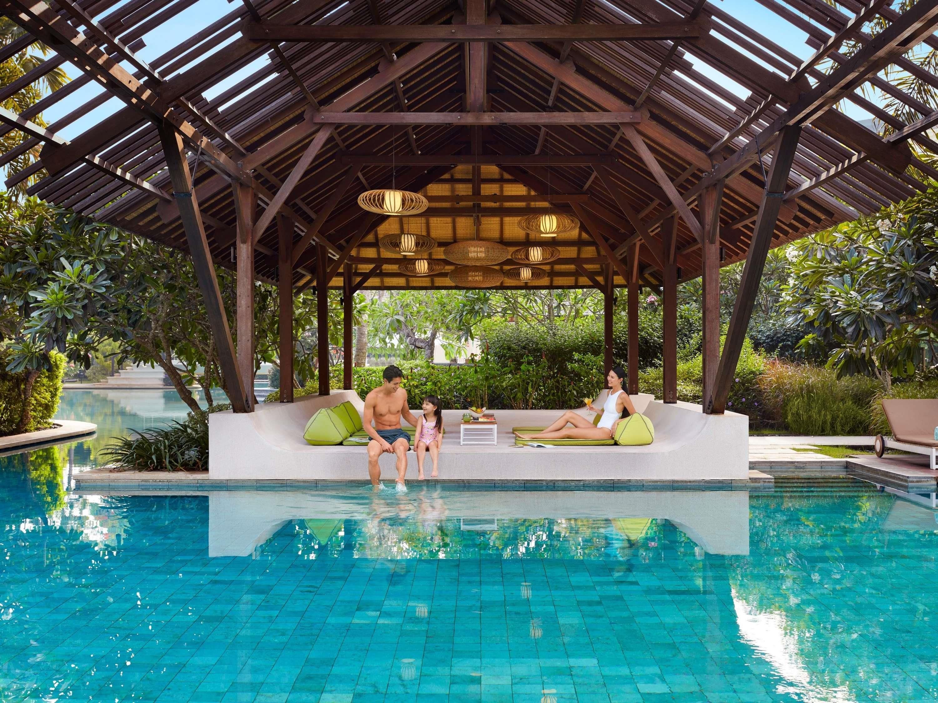 A covered pool area with a wooden pergola, lounge chairs, and a seating area with cushions.