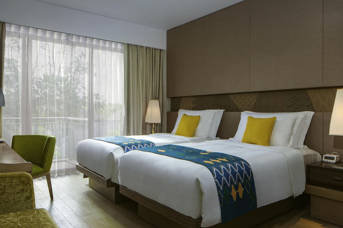 A hotel room with two beds, a green chair, a nightstand, and a window with sheer curtains.