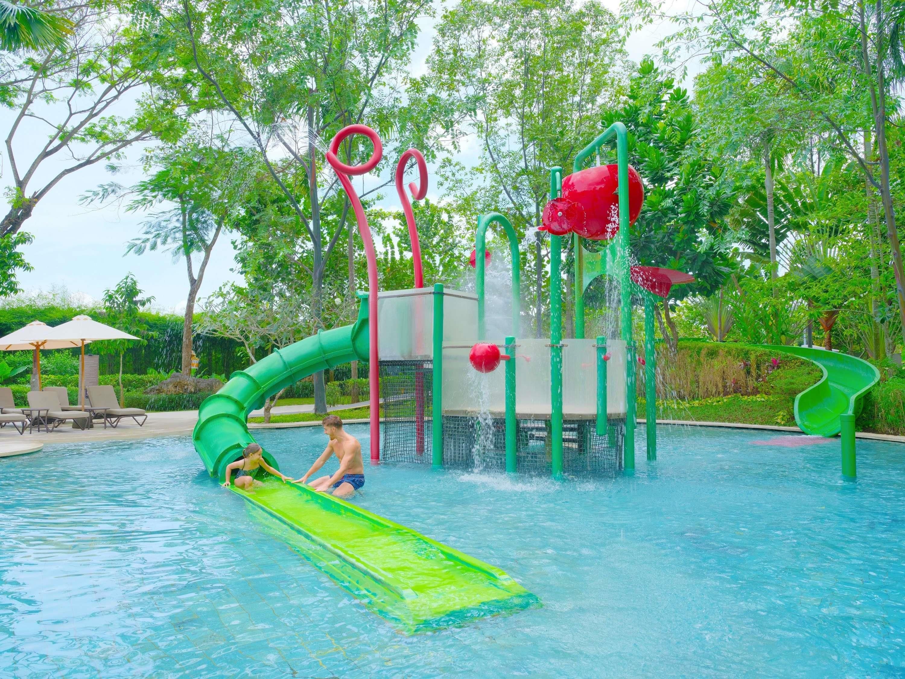 Un parque acuático con un tobogán verde, una fuente de agua roja y una piscina rodeada de árboles.