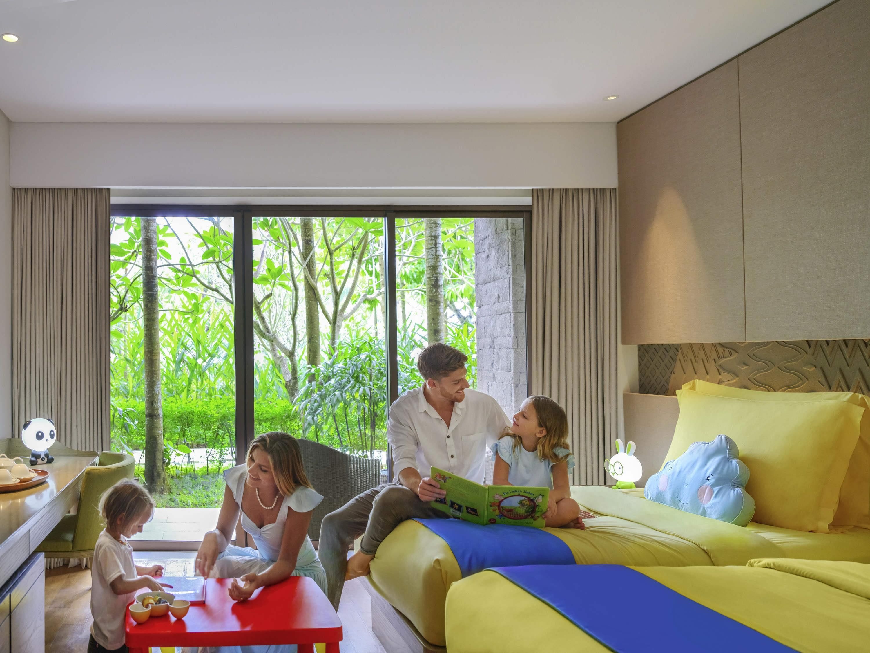 A family in a modern hotel room with a large bed, a dining table, and a view of greenery outside.