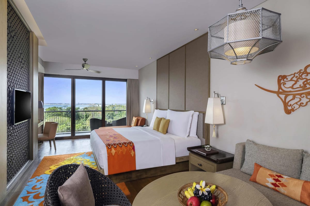 A hotel room with a bed, a TV, a balcony with a view, a dining area with a table and chairs, and a fruit basket.