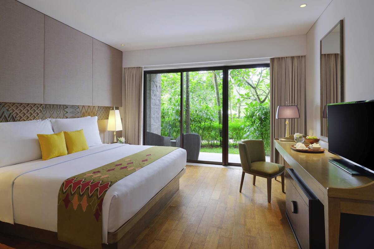 A hotel room with a large bed, a desk with a TV, and a view of greenery through the sliding glass door.
