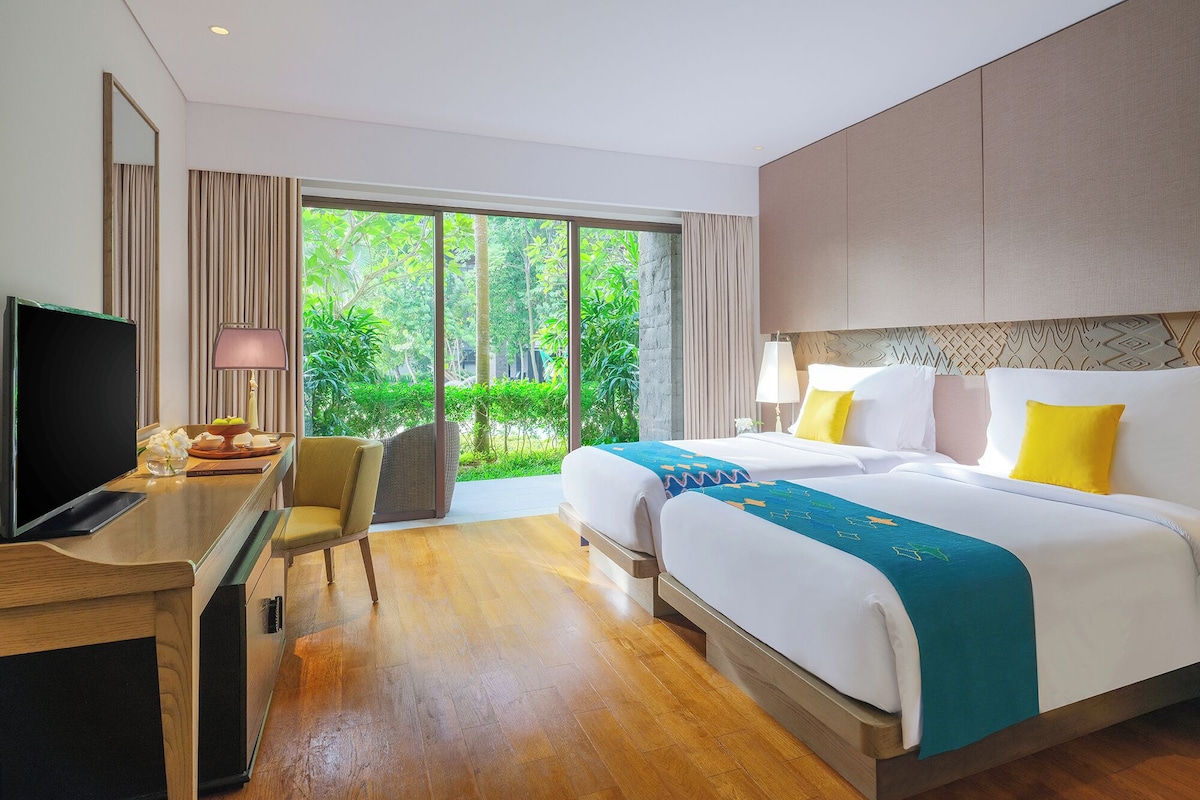 A hotel room with a large bed, a desk with a TV, and a view of greenery outside.