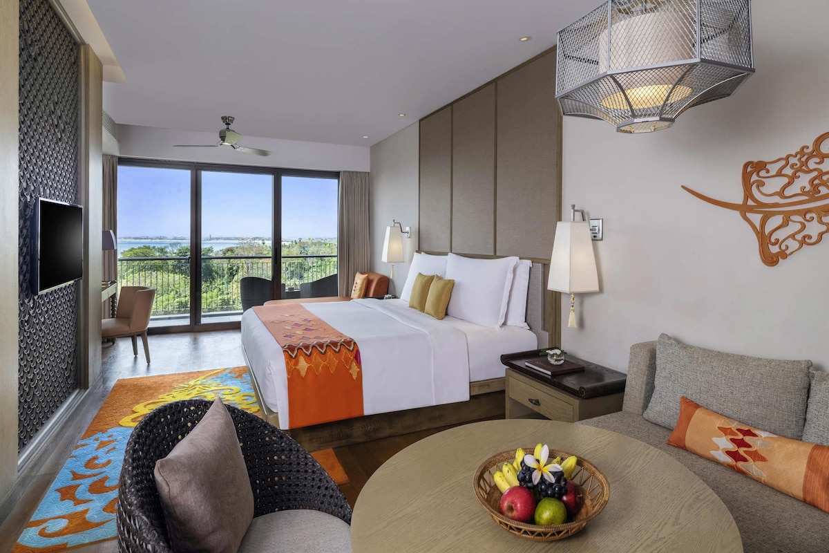 A hotel room with a bed, a TV, a balcony with a view, a dining area with a table and chairs, and a fruit basket.