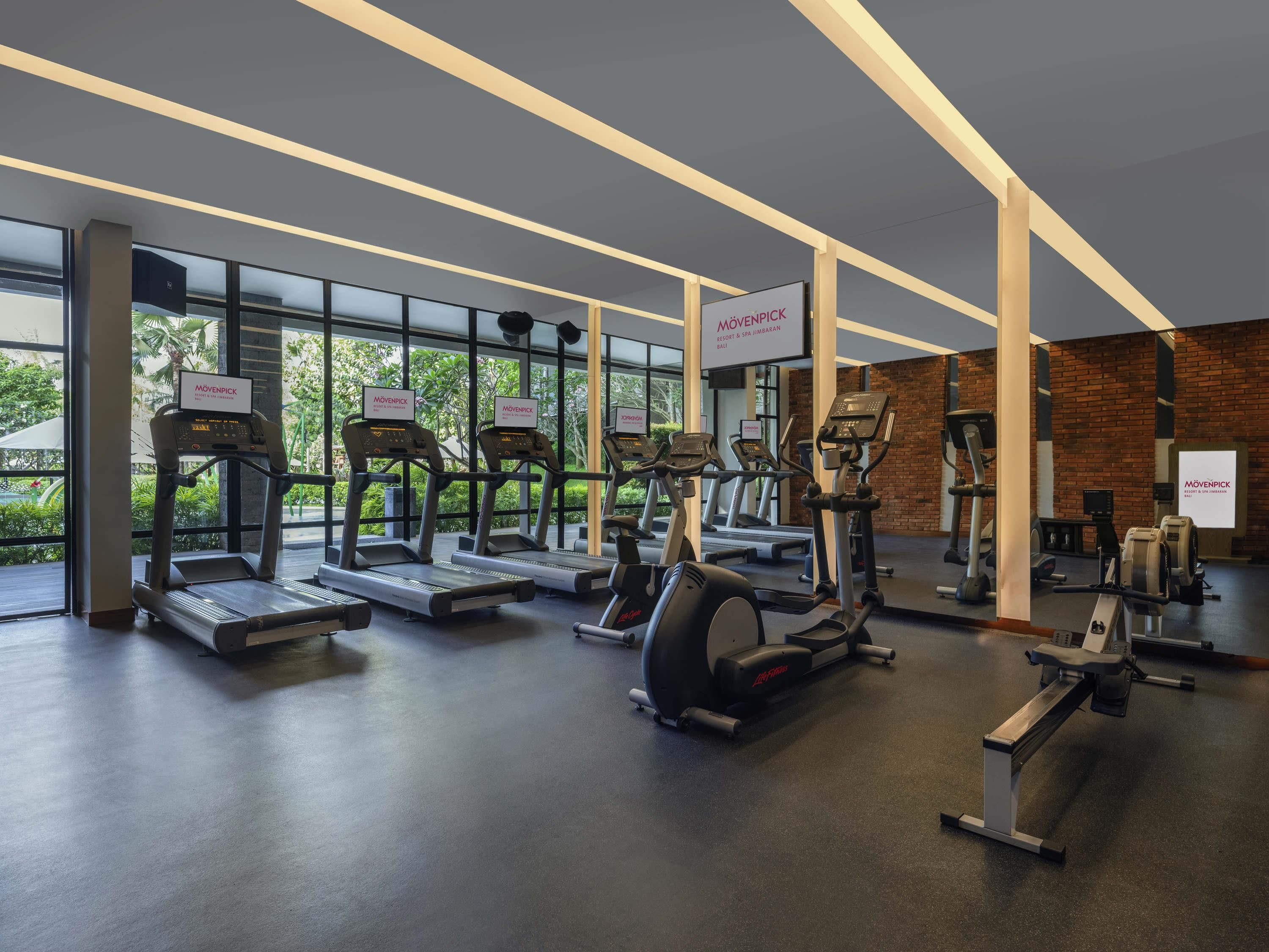 A gym with treadmills, ellipticals, and weight machines, large windows, and a brick wall.