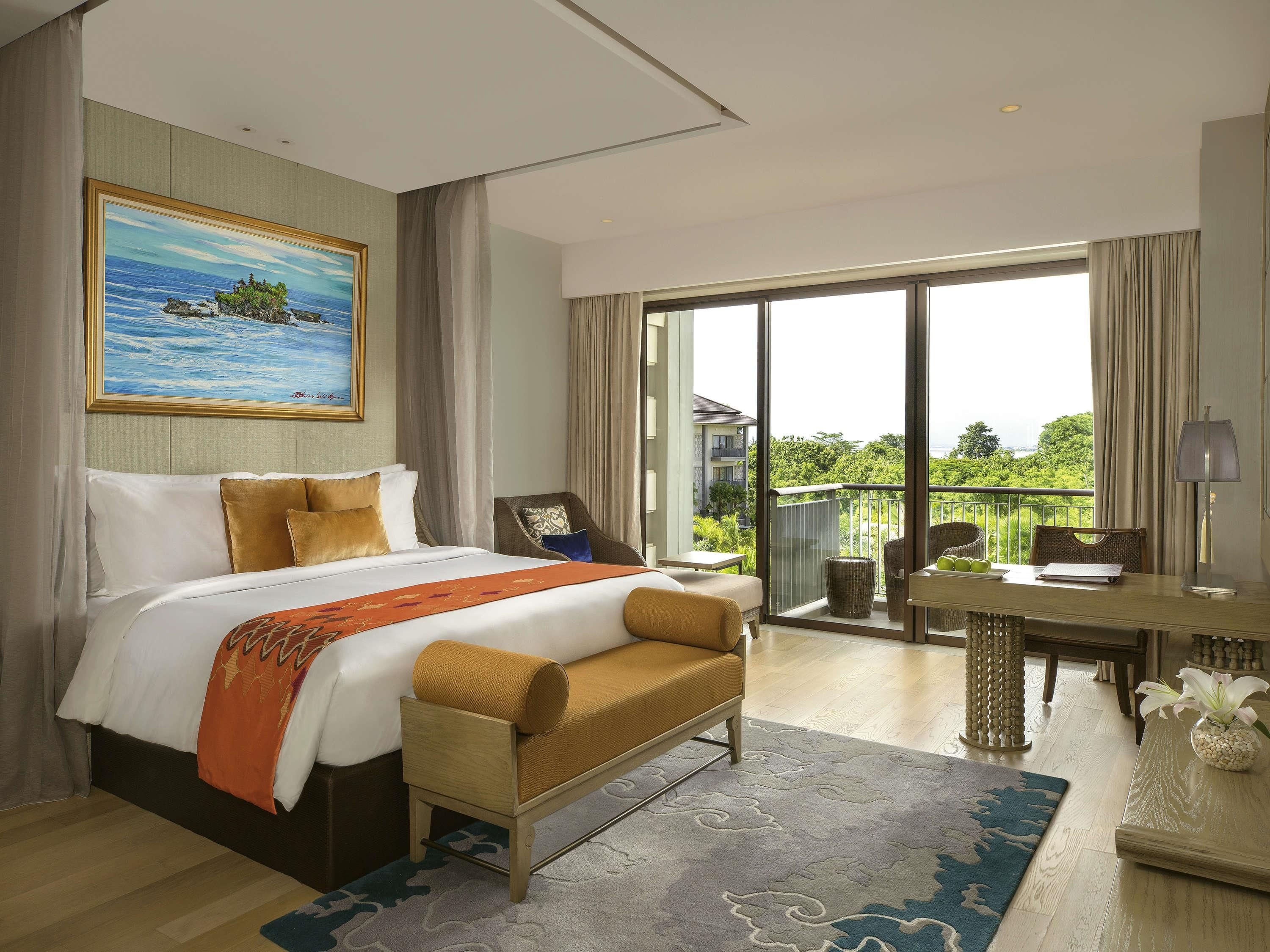 A hotel room with a bed, a desk, a chair, a sofa, and a balcony with a view of greenery.