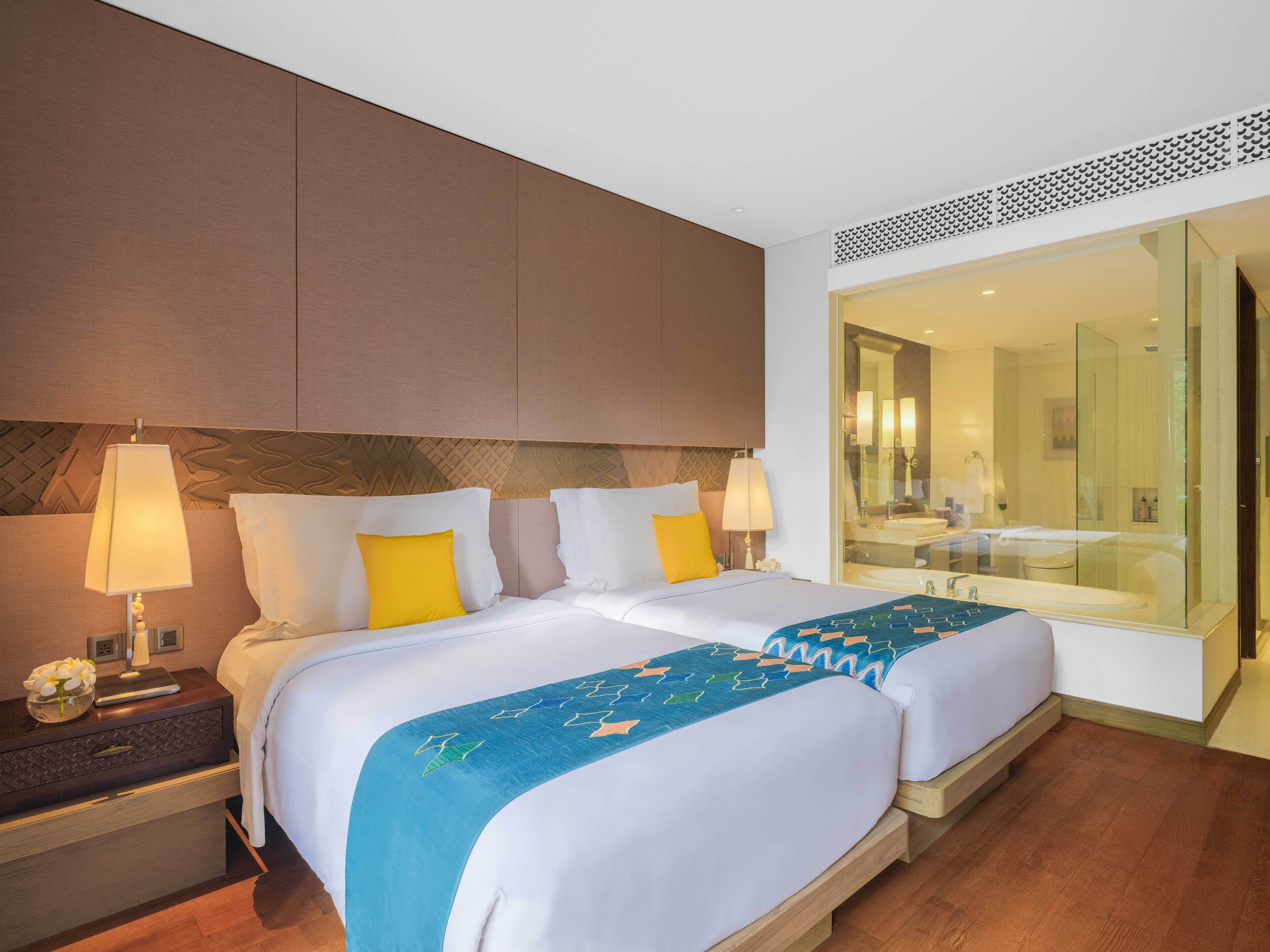 A hotel room with two beds, a nightstand, a lamp, and a large mirror reflecting a bathroom with a bathtub and sink.