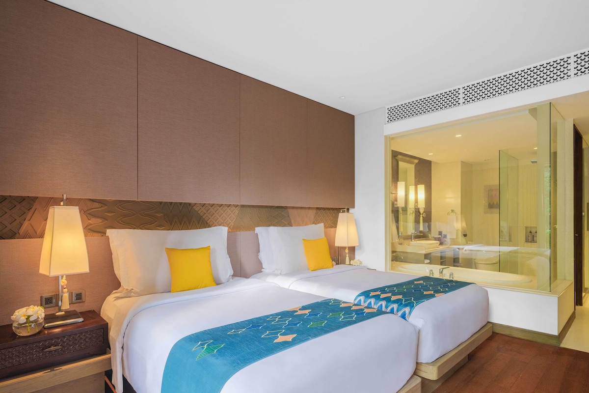 A hotel room with two beds, a nightstand, a lamp, and a large mirror reflecting a bathroom with a bathtub and sink.