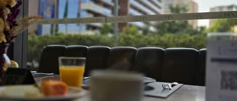 A dining table with a cup, a glass of orange juice, and a plate with food. There is a window with a view of tall buildings and greenery.
