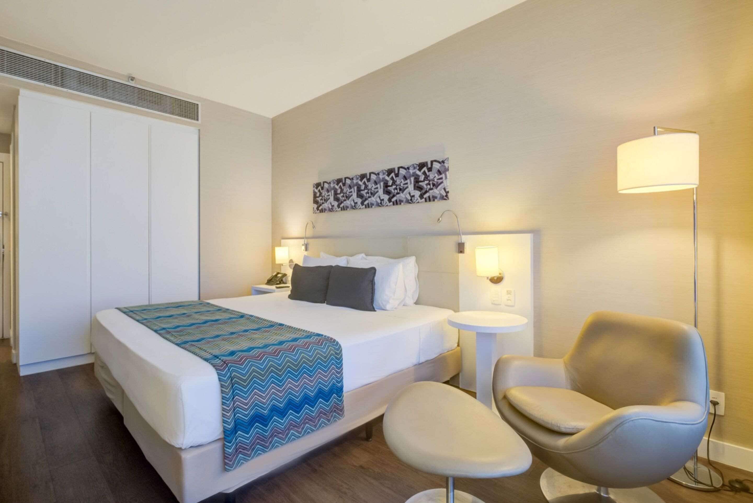 Quarto de hotel com cama, mesa de cabeceira, cadeira e obra de arte na parede.
