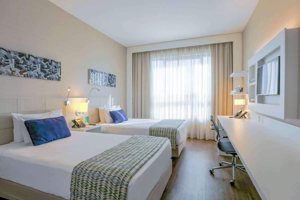 Une chambre d’hôtel avec deux lits, un bureau, une chaise, une télévision et des œuvres d’art accrochées aux murs.