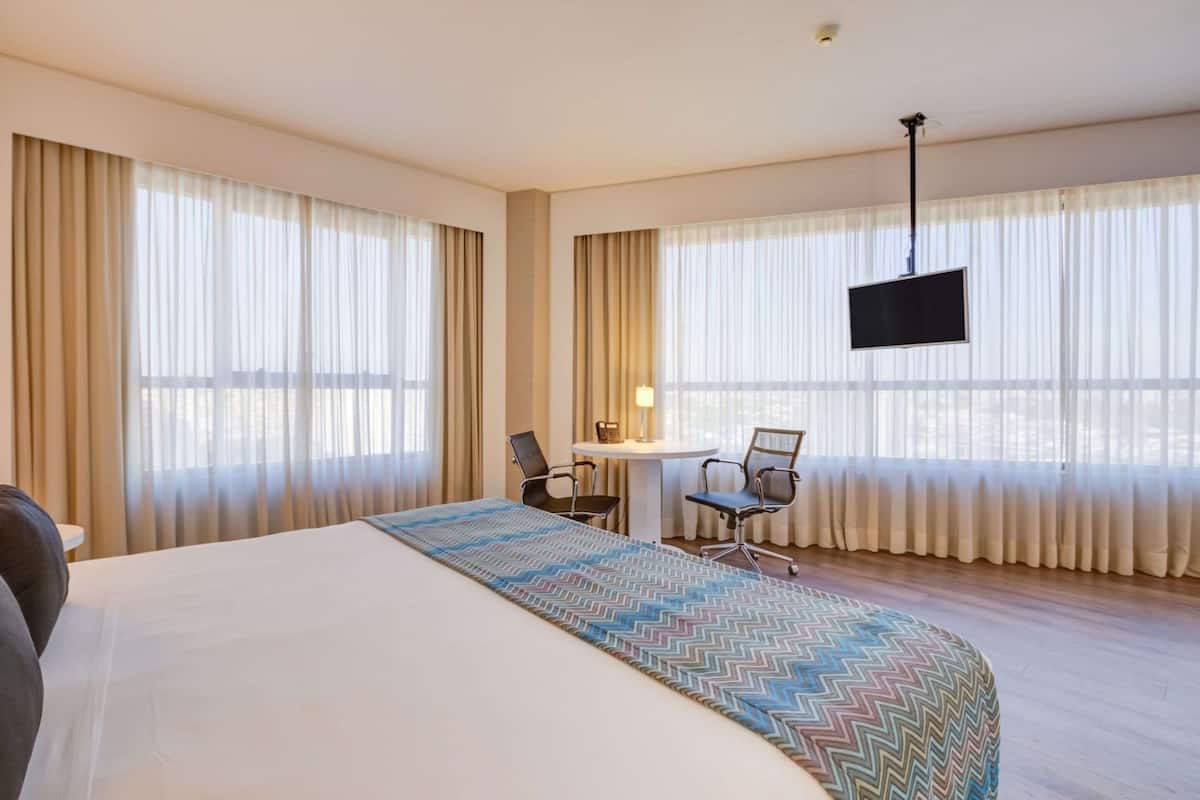 Une chambre d’hôtel avec un grand lit, un bureau avec deux chaises, un téléviseur fixé au mur et de grandes fenêtres avec des rideaux.