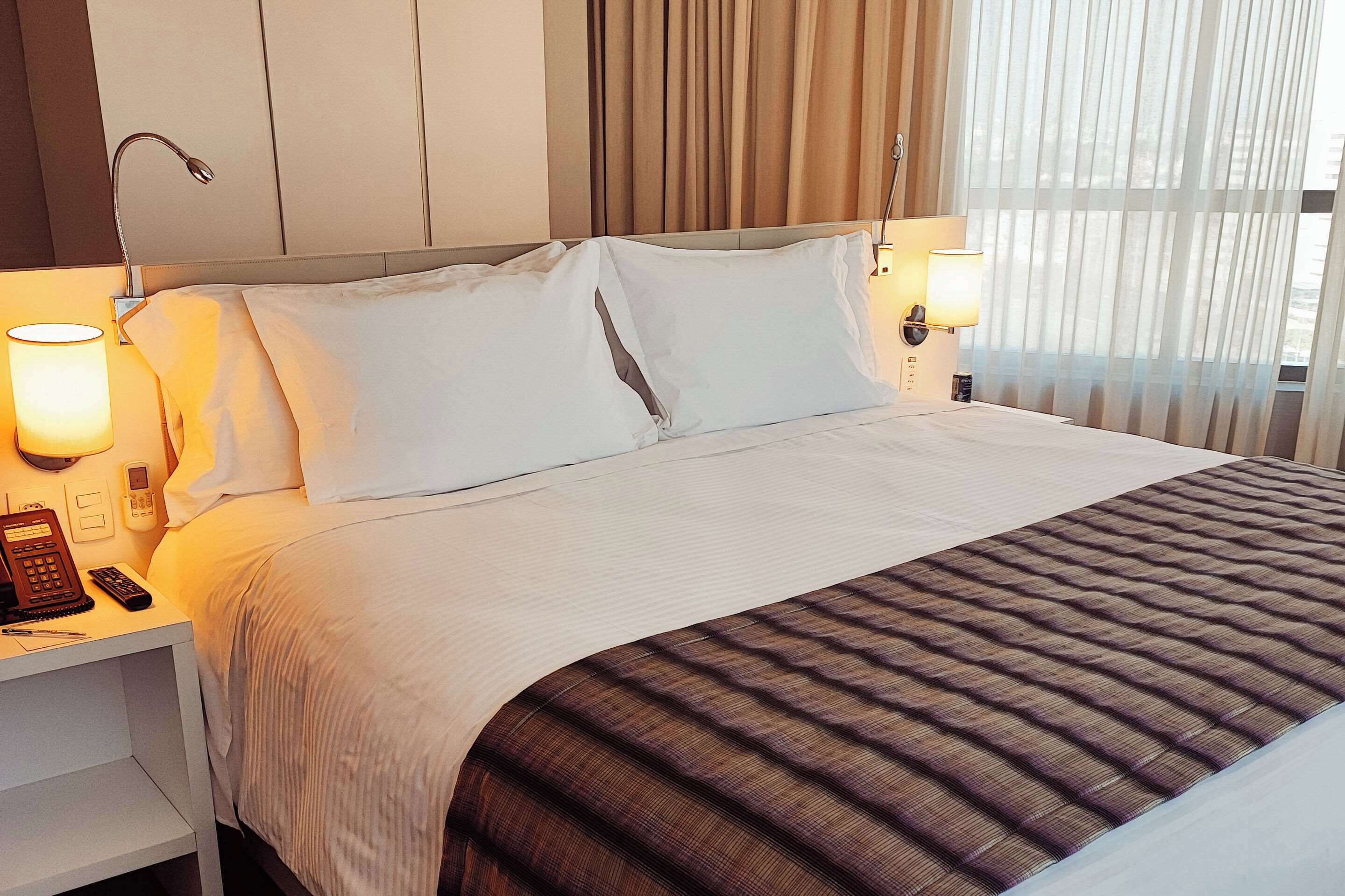 A neatly made bed with white linens and a striped blanket, bedside lamps, a telephone, and a nightstand.