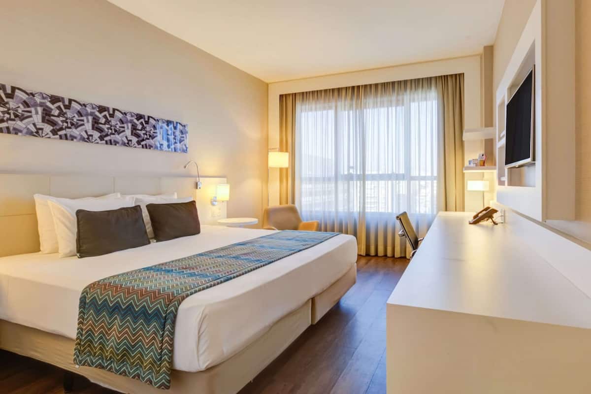 Une chambre d’hôtel avec un grand lit, un bureau, une chaise, une télévision et une fenêtre avec des rideaux.