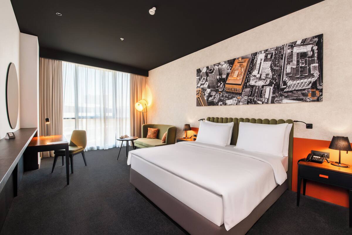 Une chambre d’hôtel avec un grand lit, un bureau, une chaise, un canapé et un tableau représentant un paysage urbain accroché au mur.
