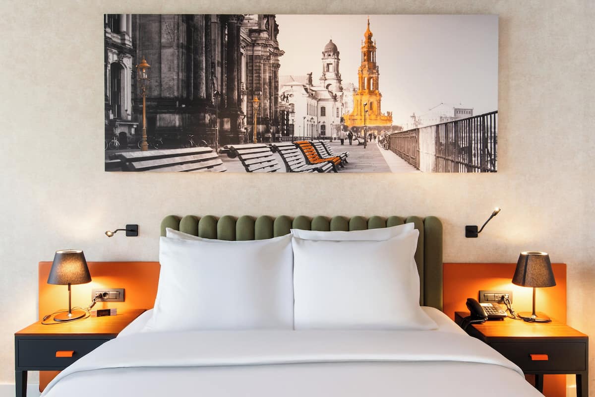 Une chambre d’hôtel avec une grande œuvre d’art représentant un paysage urbain, encadrée, une tête de lit recouverte d’un tissu vert, du linge de lit blanc et deux tables de chevet avec des lampes et un téléphone.