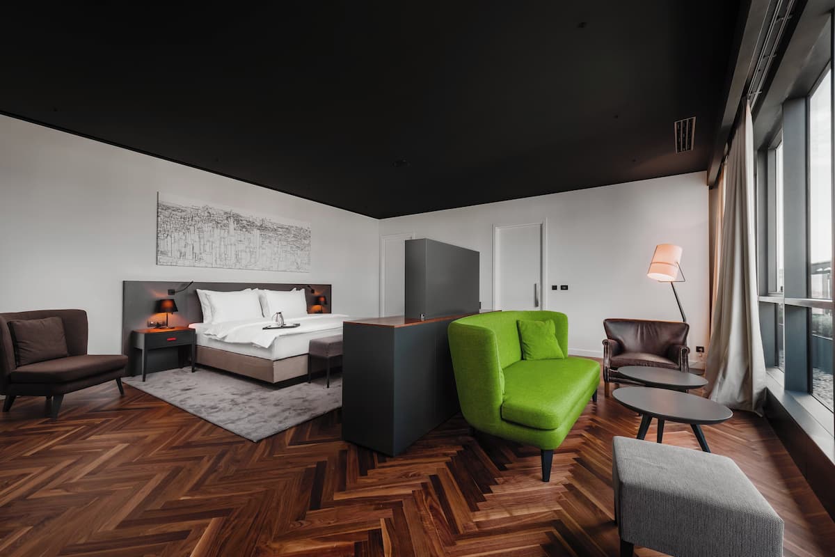 Une chambre d’hôtel moderne avec un lit, un fauteuil vert, un fauteuil marron, une petite table et une lampe.