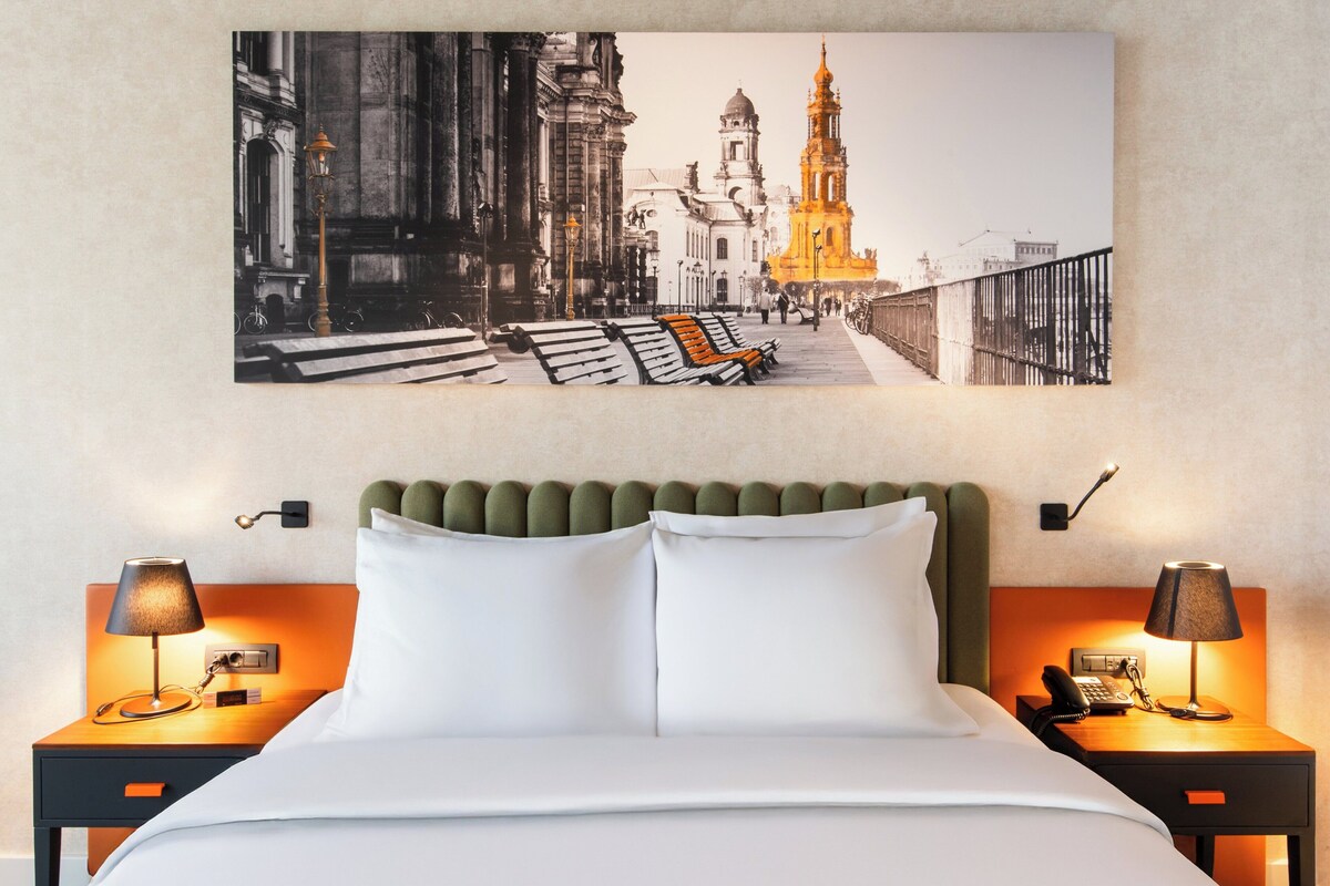 Une chambre d’hôtel avec une grande œuvre d’art représentant un paysage urbain, encadrée, une tête de lit recouverte d’un tissu vert, du linge de lit blanc et deux tables de chevet avec des lampes et un téléphone.