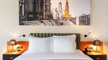 A hotel room with a large framed cityscape artwork, a green upholstered headboard, white bedding, and two bedside tables with lamps and a phone.